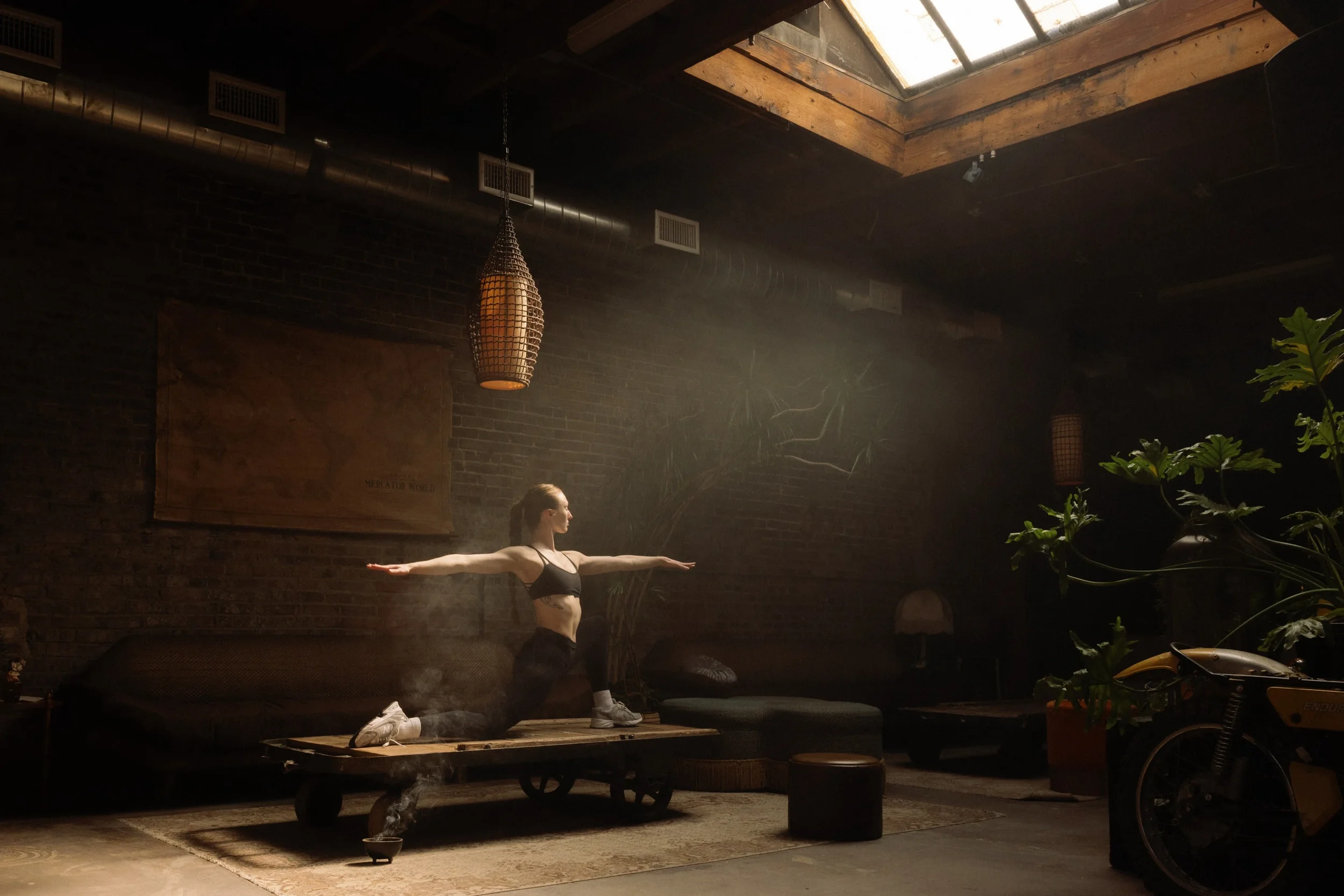 A woman in black athletic wear doing yoga on a wooden platform in a dimly lit room with brick walls, hanging lanterns, and a skylight.