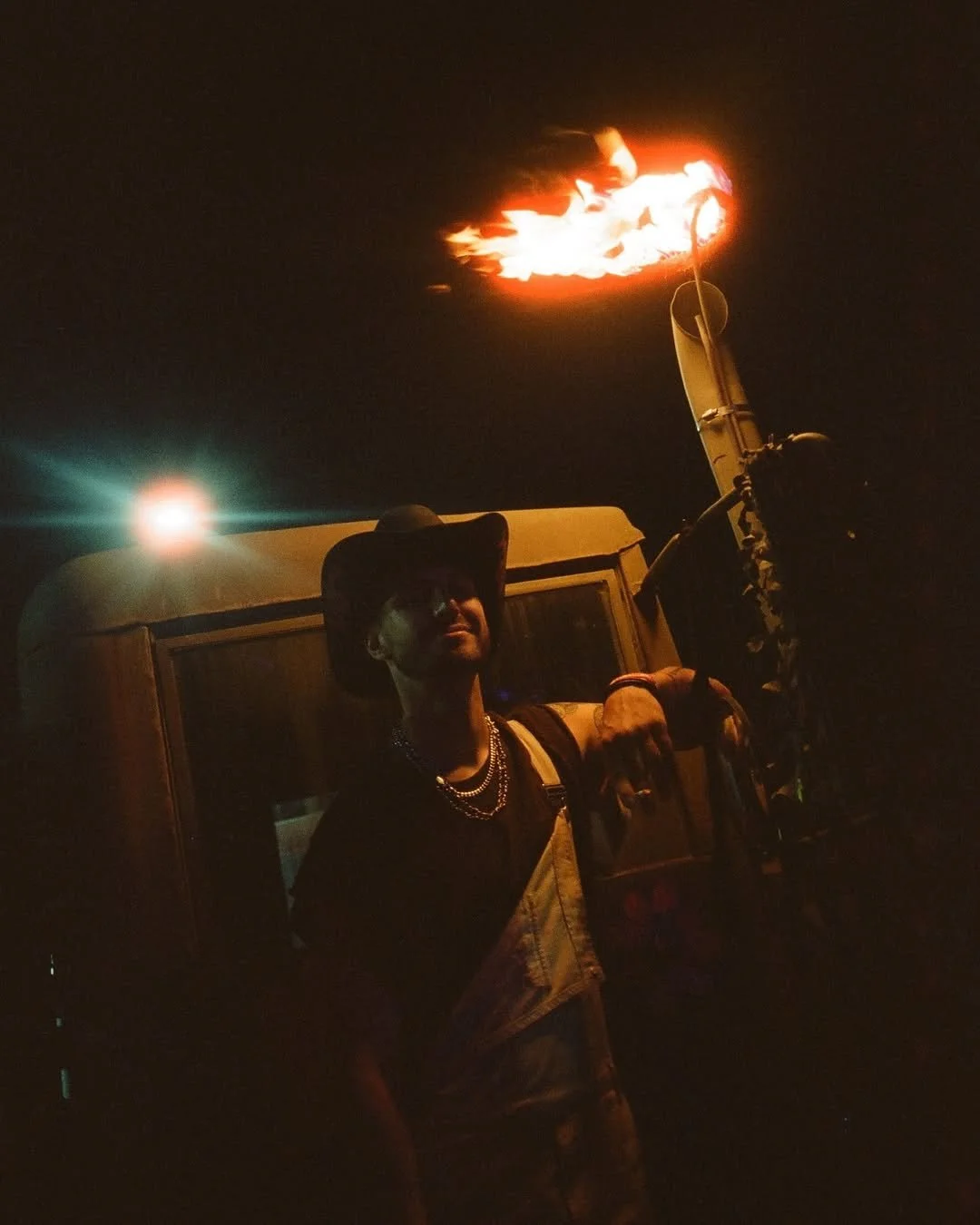 A person in sunglasses, a cowboy hat, and chains standing next to a vehicle at night, holding a flame-throwing device that is emitting a large flame into the dark sky.