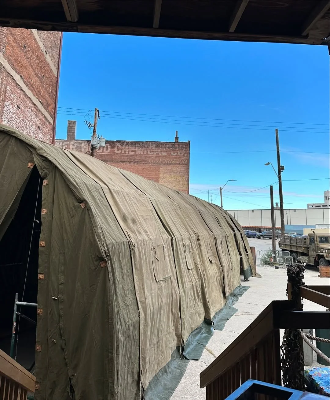 A military-style beige tent set up outdoors, with a brick building and a parking lot in the background under a clear blue sky.