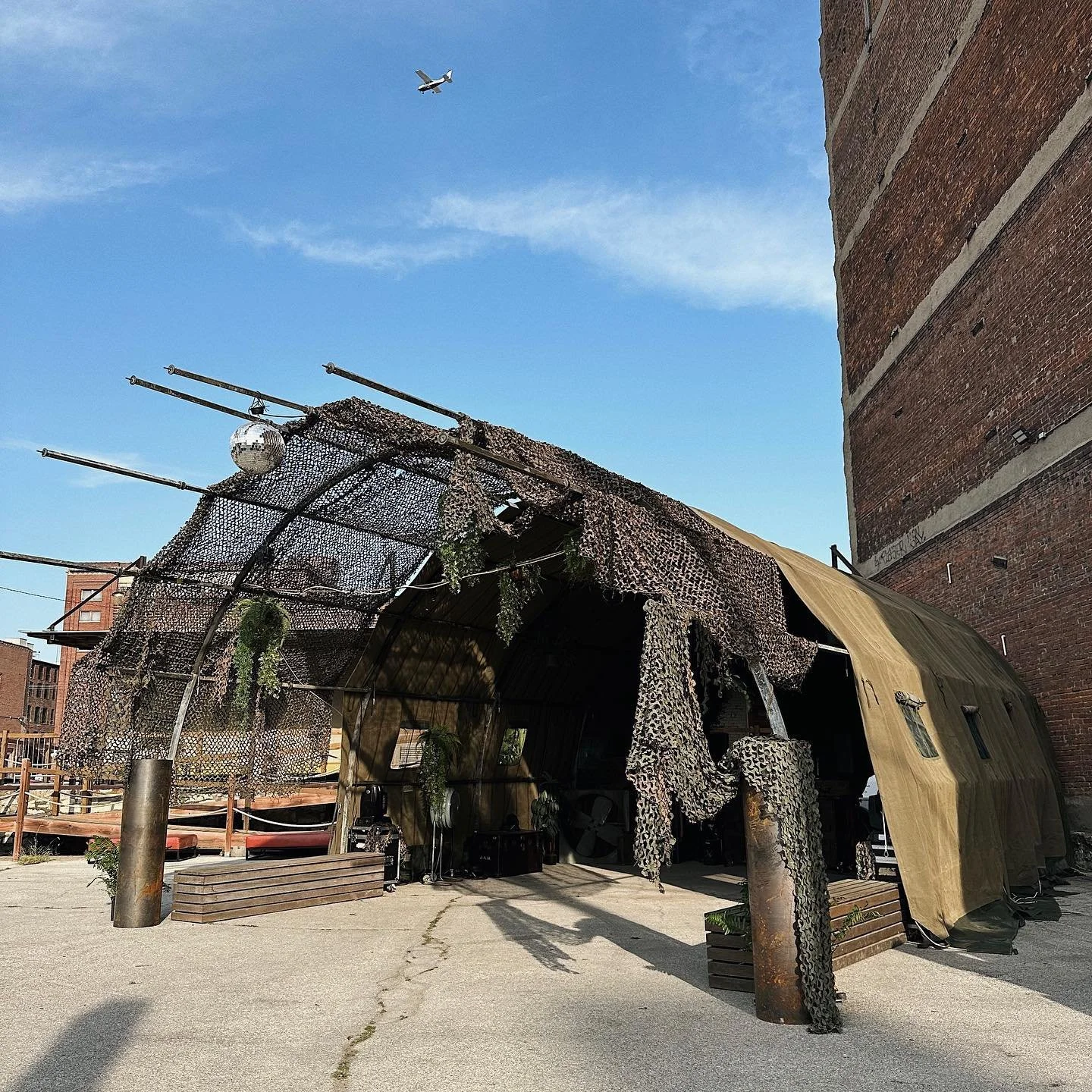 A makeshift outdoor shelter made of camouflage netting and a beige tarp next to a brick wall, with a large cylindrical metal post and green plants hanging from it. An airplane is flying in the blue sky above.