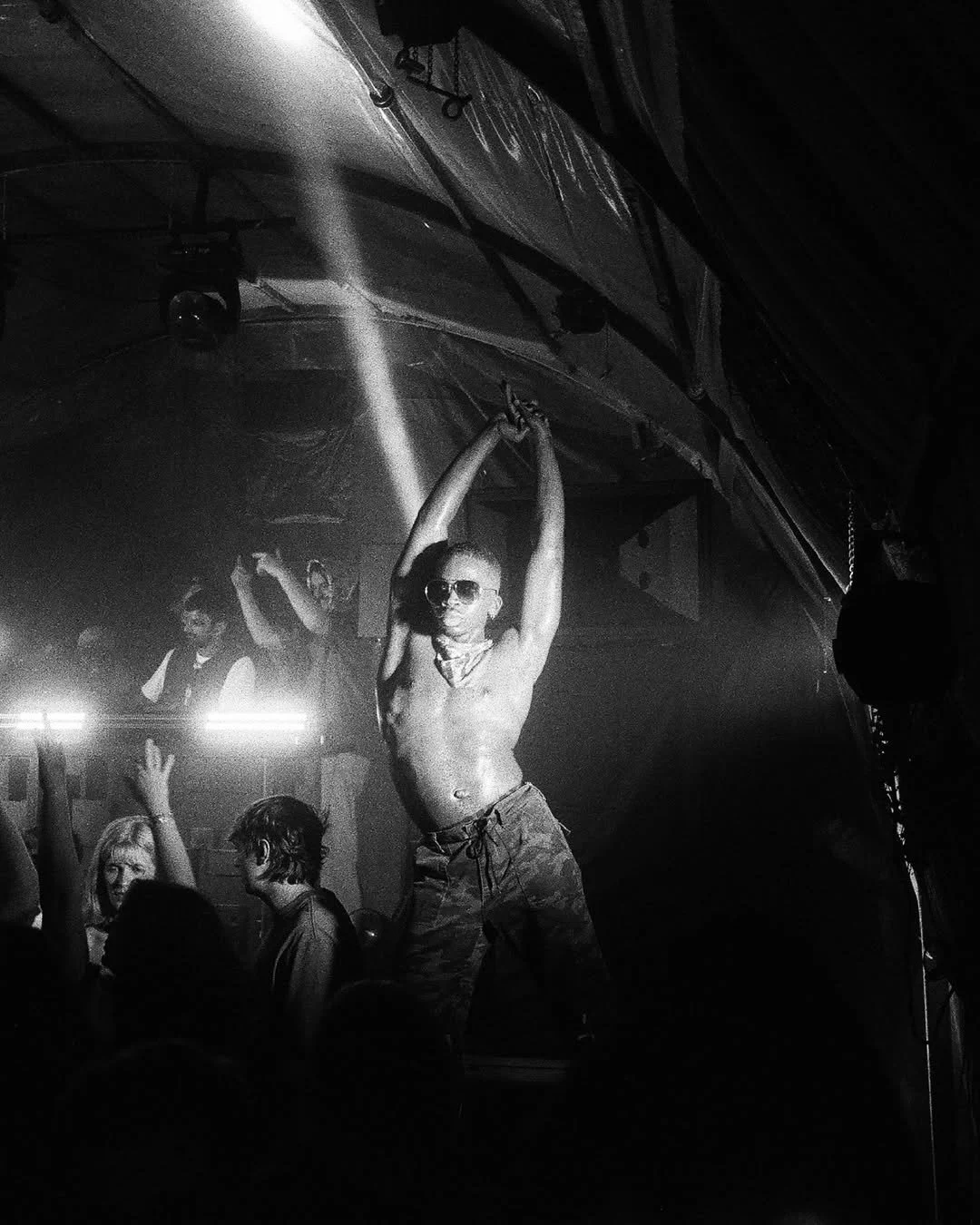 A shirtless man with sunglasses is dancing on stage with his arms raised above his head. Several people are dancing around him in a nightclub or concert setting with dark lighting and a crowd in the background.