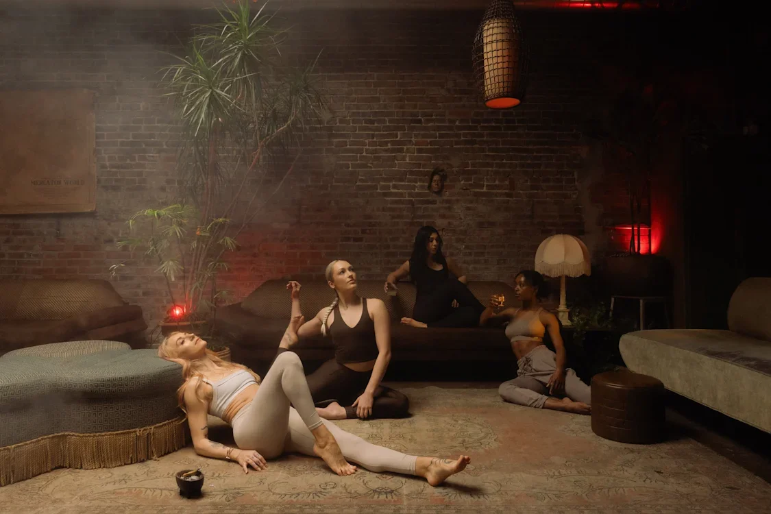 Four women in a dimly lit room with brick walls, some sitting or lying down, practicing yoga or stretching, with a large plant, a carpet, and vintage decor including a lamp and a hanging light.