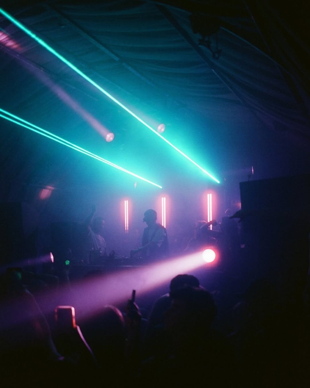 A dark nightclub scene with DJs on stage illuminated by colorful laser lights and vertical LED lights, with silhouettes of people in the foreground.