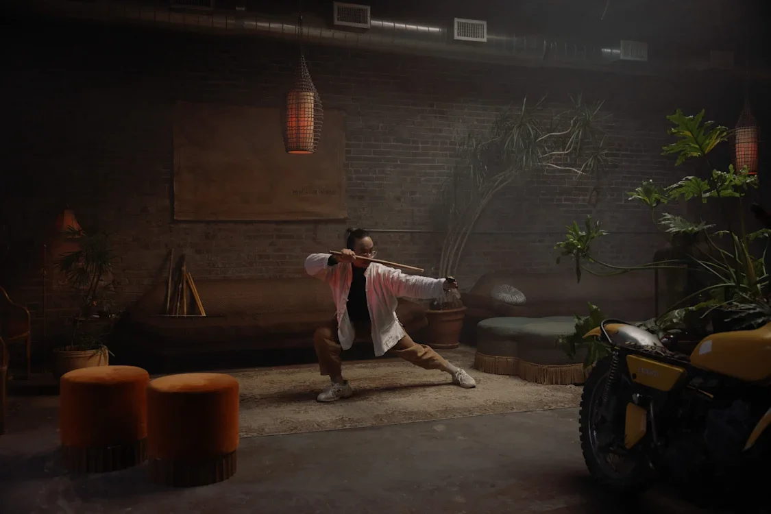Person practicing martial arts with a sword indoors, surrounded by plants, furniture, and a motorcycle