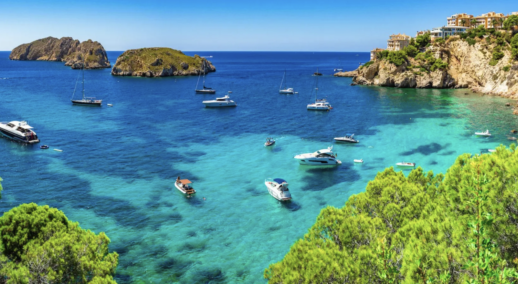 Clear blue water with several boats and yachts anchored near rocky cliffs and a hillside with luxurious houses, surrounded by green trees.