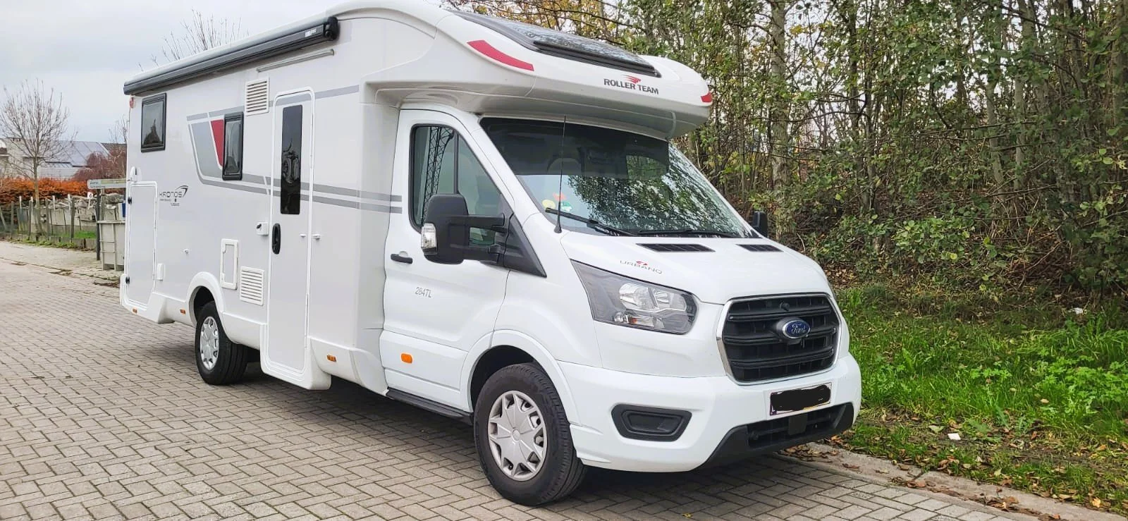 Wit camper op een oprit met groene struiken en bomen op de achtergrond.