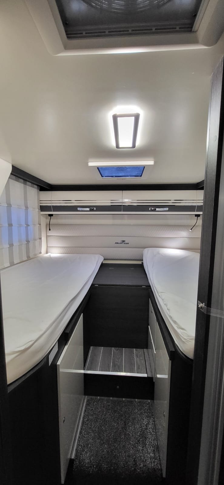 Binnenkant van een camper of caravan met twee bedden, een plafondlamp, een dakraam en een bedrand met ingebouwde lampjes.