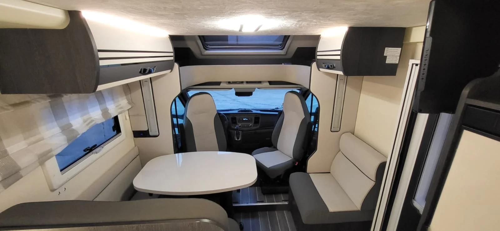 Interieur van een camper of buscampingvoertuig, met twee bestuurdersstoelen, een eethoek met bank en tafel, en ramen.
