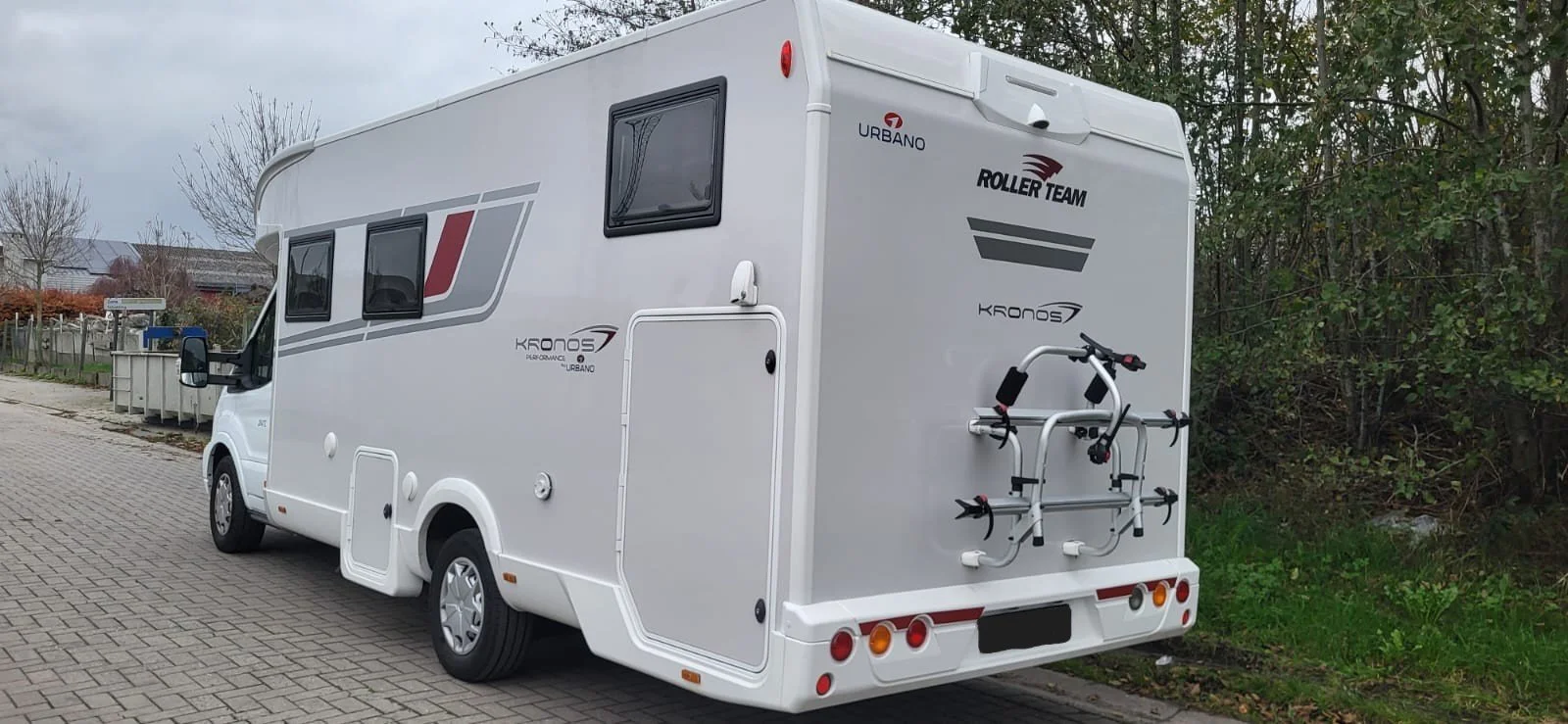 Wit camper met fietsendrager, parkt op een straat met bomen op de achtergrond.