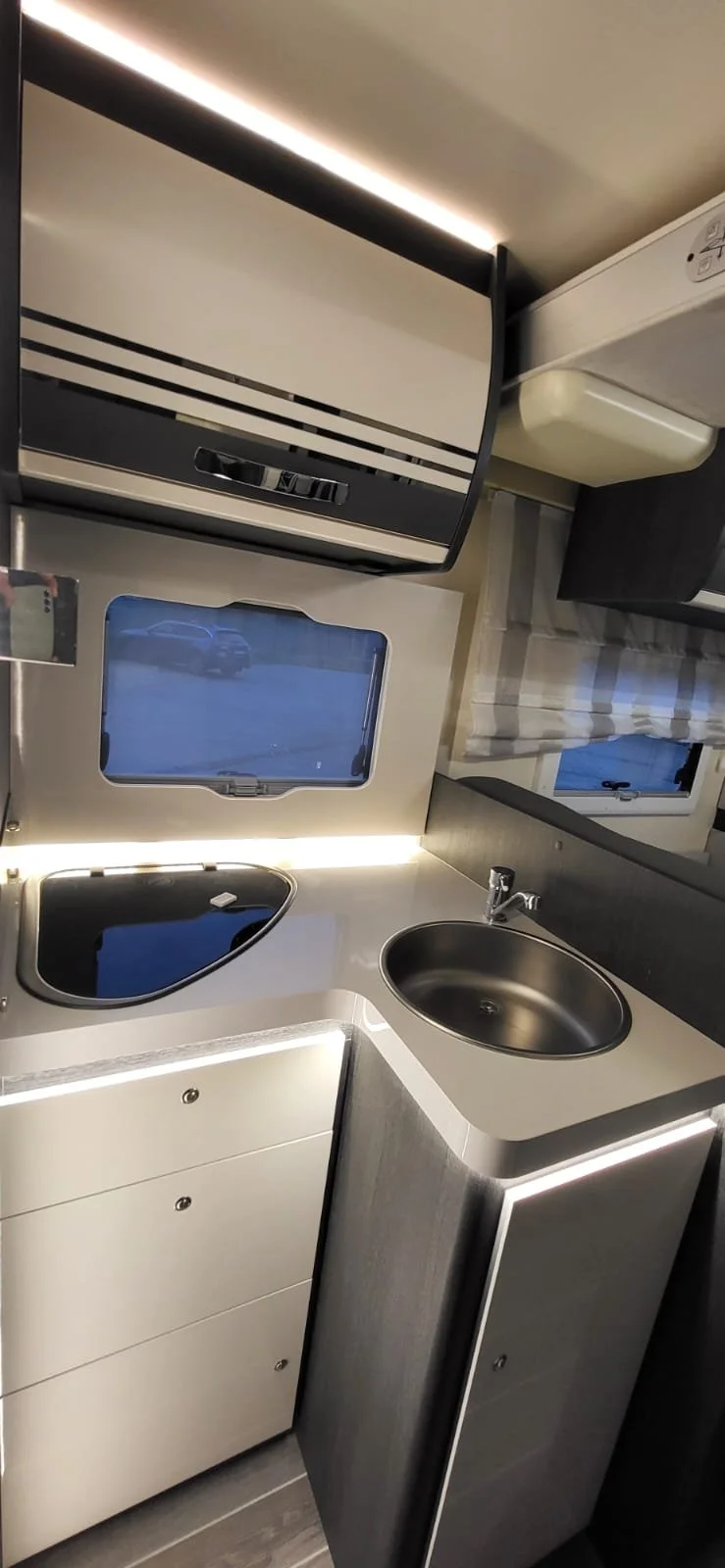 De afbeelding toont de binnenkant van een kleine keukenruimte in een camper of caravan, met een ronde spoelbak, een klein raam en kasten. Er liggen ook horren en een lamp boven de keuken.