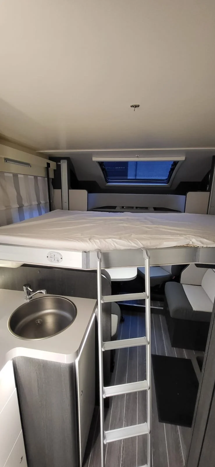 Binnenkant van een camper of kleinen room met een hoogslaper, een luik op het plafond en een kleine gootsteen, met stoel en tafel eronder.