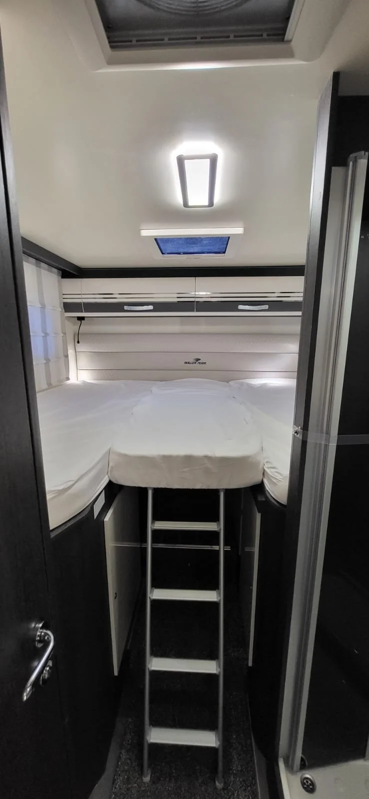 Een compact slaapkamers in een camper of kleine ruimte, met een tweepersoonsbed en een ladder die naar het bed leidt. Boven het bed zijn ramen en plafondverlichting.