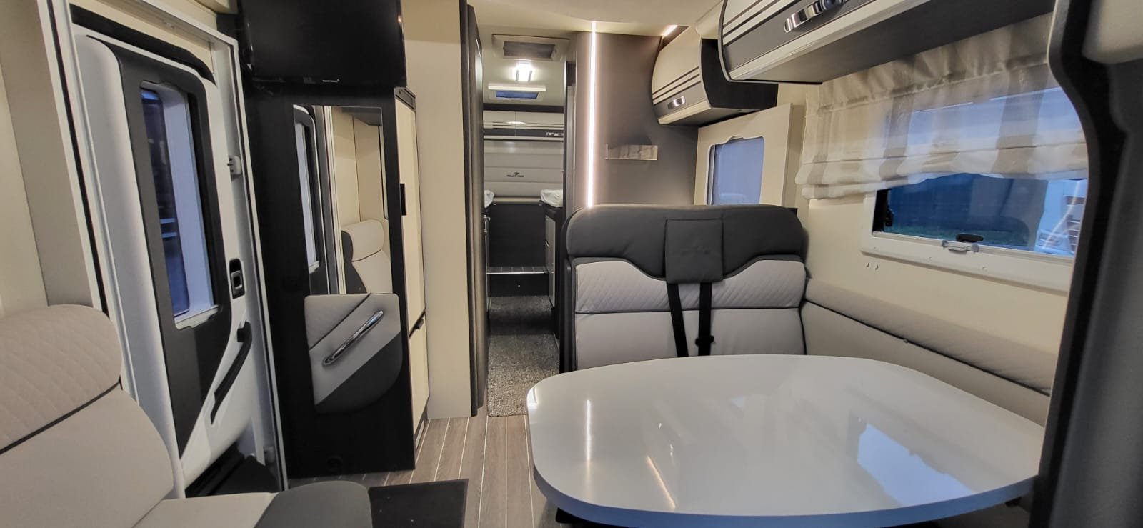 Interieur van een camper of recreatievoertuig met zithoek, tafel, ramen, en kitchenette in de achtergrond.