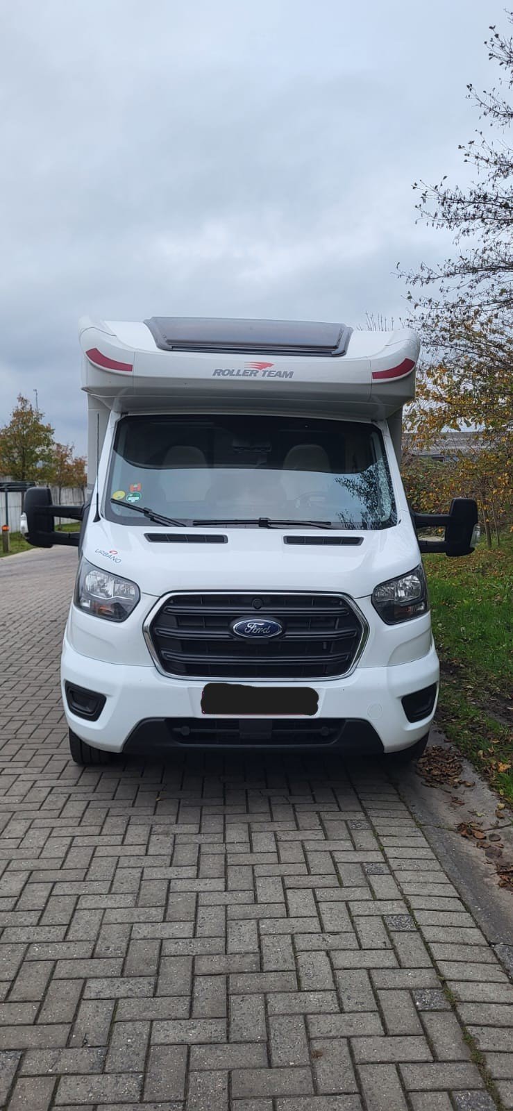 Wit camperwagen met 'ROLLER TEAM' op de bovenkant geparkeerd op een stenen parkeerplaats, met herfstbomen op de achtergrond en een bewolkte lucht.