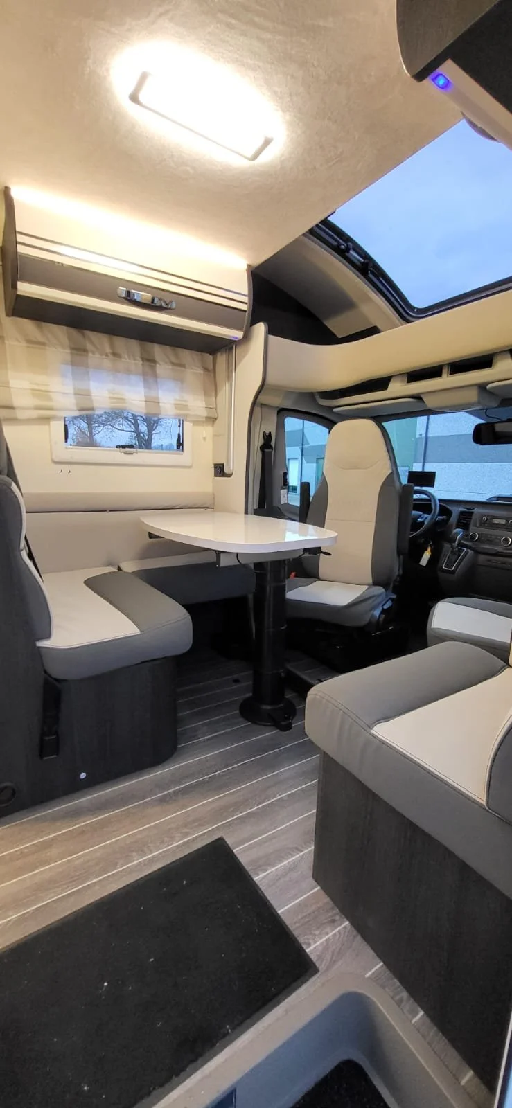 Het interieur van een campers of busje met een zit- en eethoek, een raam met een gordijn, een kast boven het raam, een tafel, en een zonnedak dat open staat.