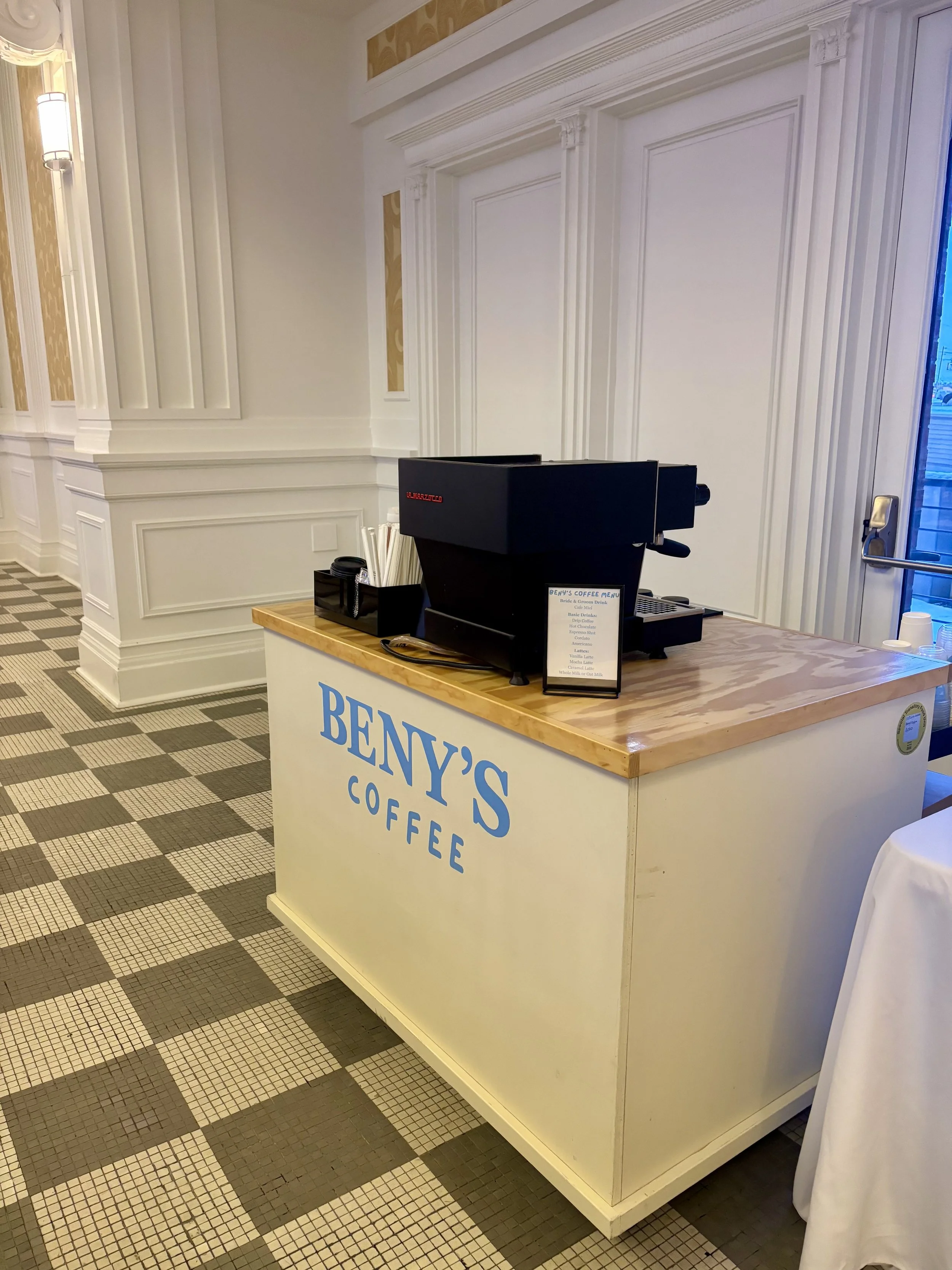 Coffee stand with a black coffee machine on a wooden countertop in an elegant, well-lit room. The stand has 'Beny's Coffee' written in blue letters on the front.