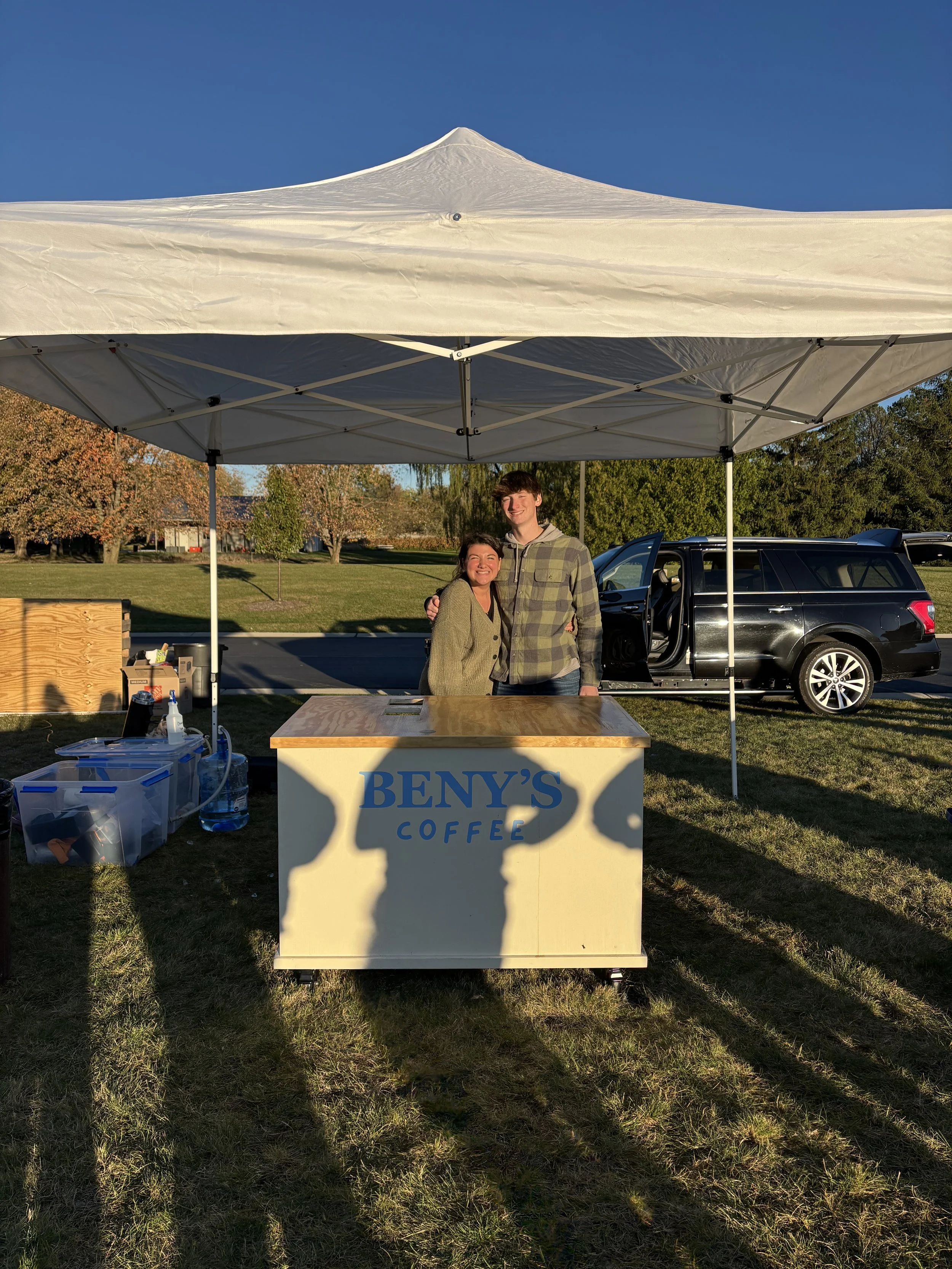 Two people standing under a white canopy tent at a booth labeled BENY'S COFFEE outdoors, with a black car, trees, and a clear blue sky in the background.