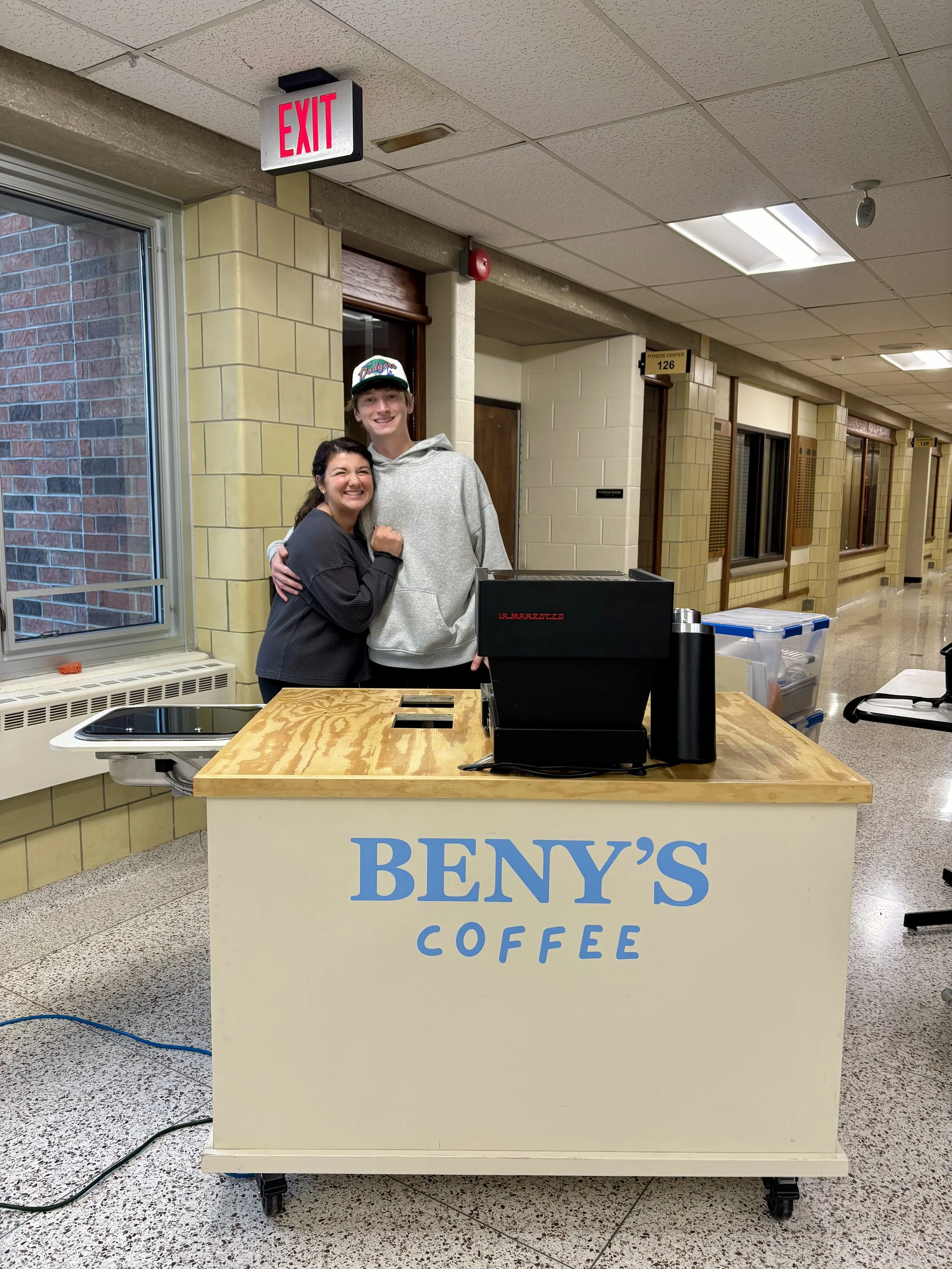 Two people, a woman and a tall man, stand together behind a coffee stand labeled 'Beny's Coffee' in a hallway, smiling with their arms around each other.