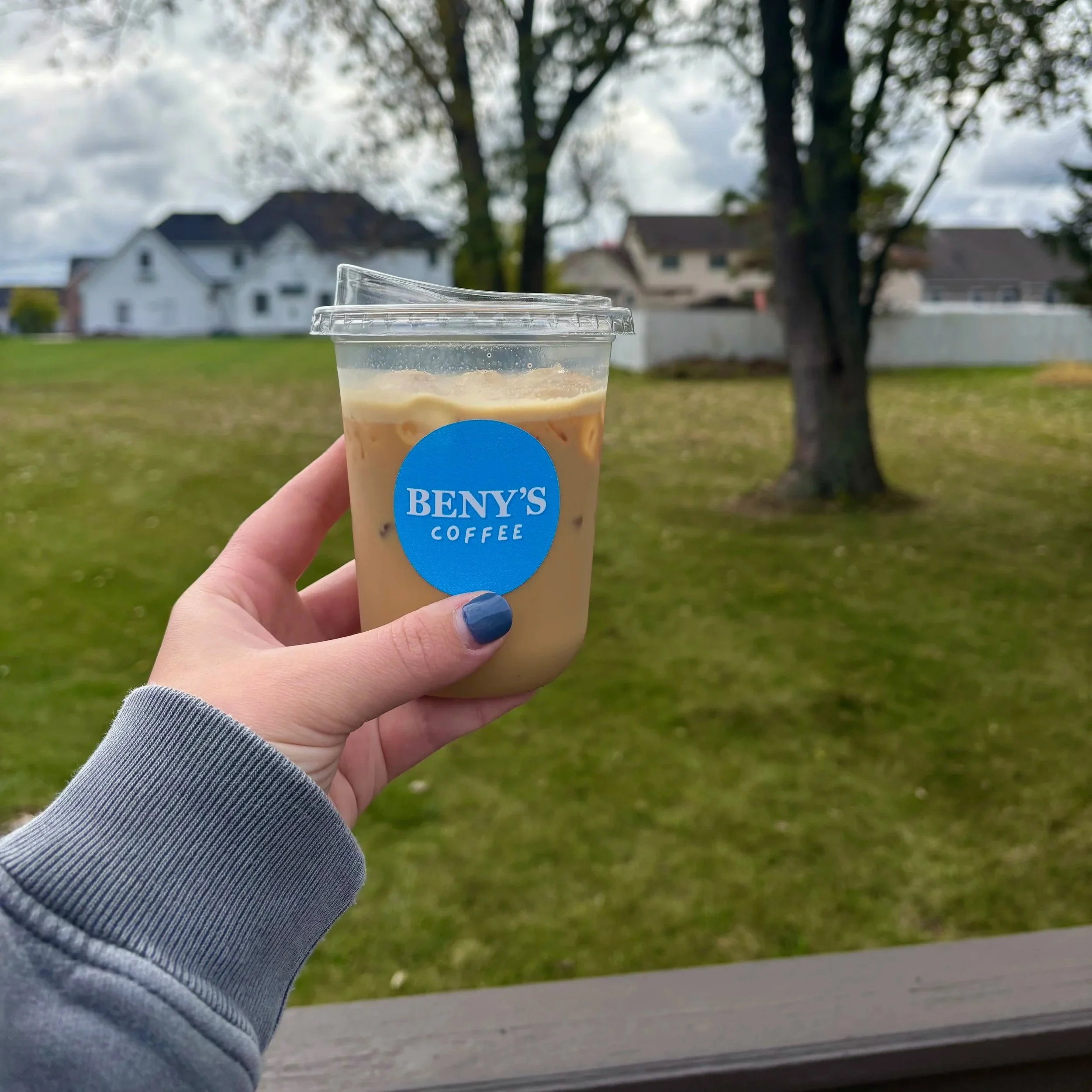 Person holding an iced coffee in a clear plastic cup with a blue Beny's Coffee logo. The person has painted nails and is wearing a gray sleeve. The background shows a grassy area with trees and houses under cloudy skies.