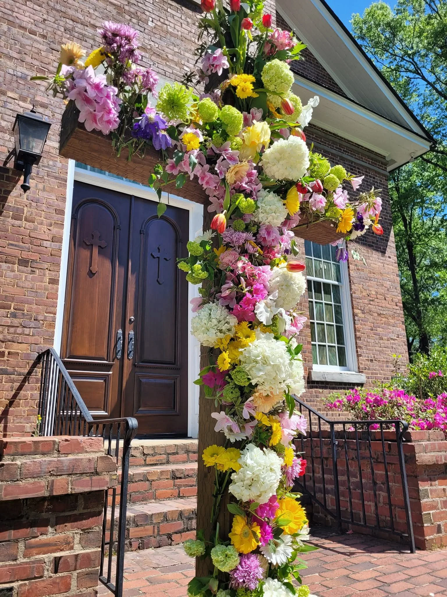Flower arrangement on a wooden pole outside a brick house with a dark wooden door and white window, surrounded by greenery and a pink flowering bush.
