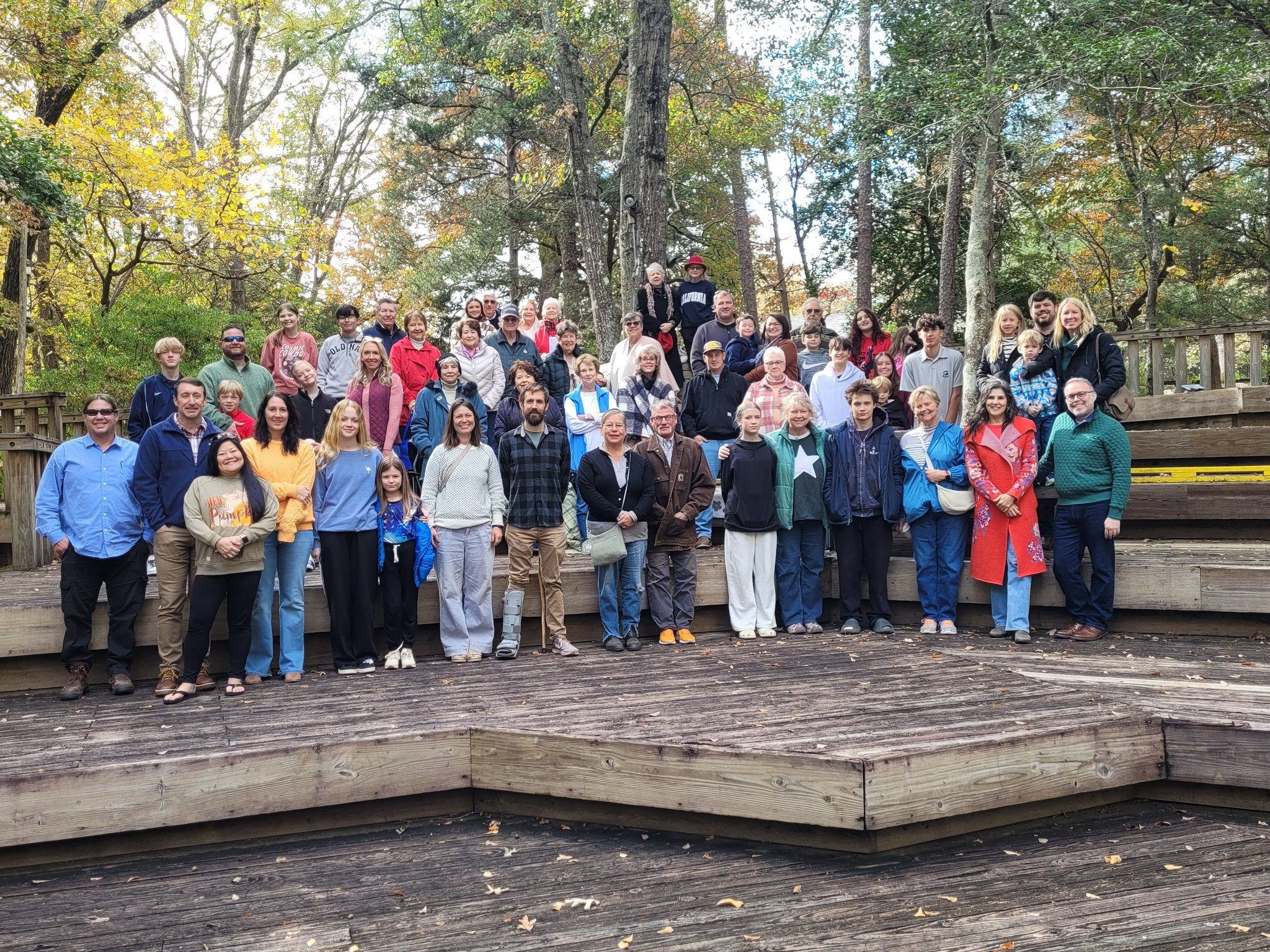 Group of people posing for a group photo outdoors on a wooden platform surrounded by trees with autumn leaves.