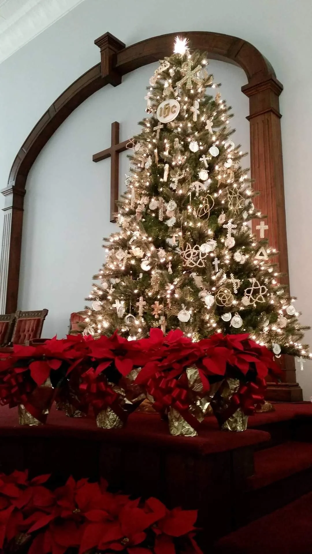 A decorated Christmas tree with white and gold ornaments and lights, set against a blue wall with a wooden cross and arch. Red poinsettia flowers and gold-wrapped gifts are at the base.