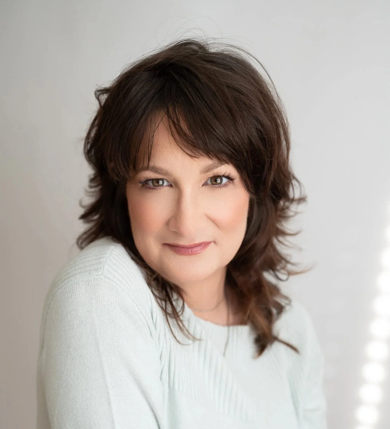 A woman with short, wavy dark brown hair and light makeup, smiling softly in front of a light gray background.