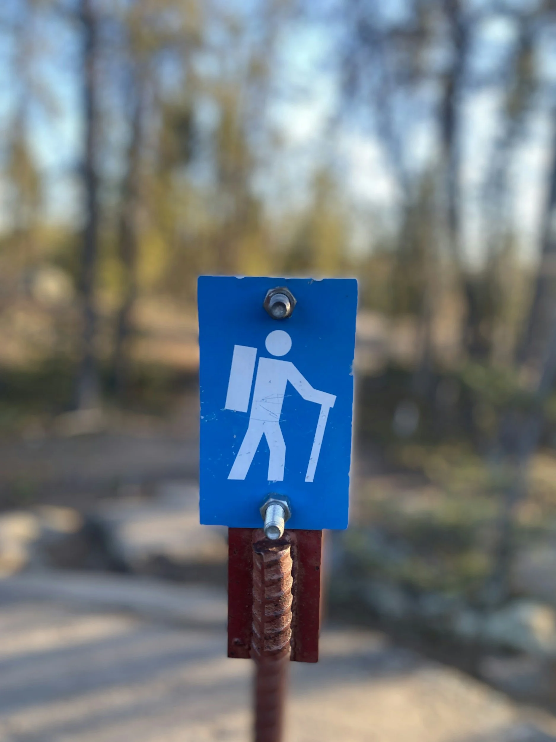 A hiking trail sign with a blue background and a white pictogram of a person with a backpack and walking stick, mounted on a rusty metal support with a nut and bolt.
