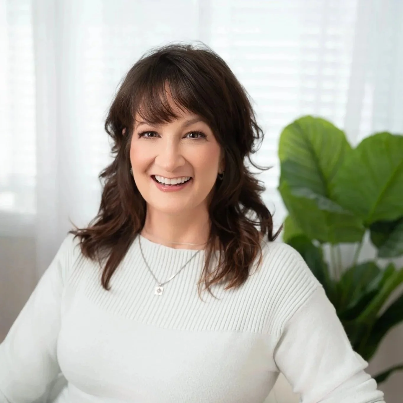 A smiling woman with dark brown, shoulder-length hair, wearing a white sweater and a necklace, sitting indoors near a green leafy plant.