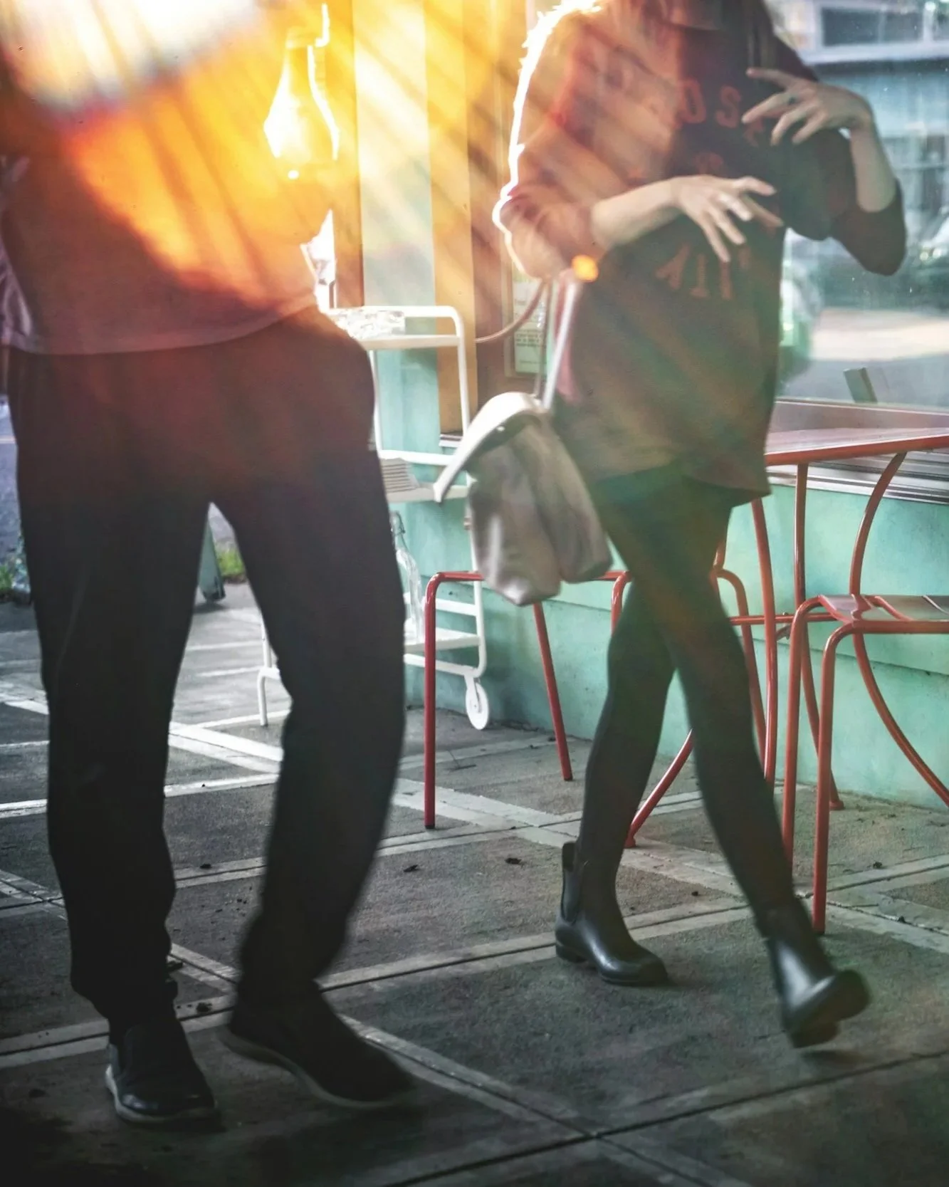 Two people standing and walking outside a shop, with sunlight streaming through the window, and an empty red chair and tables visible.