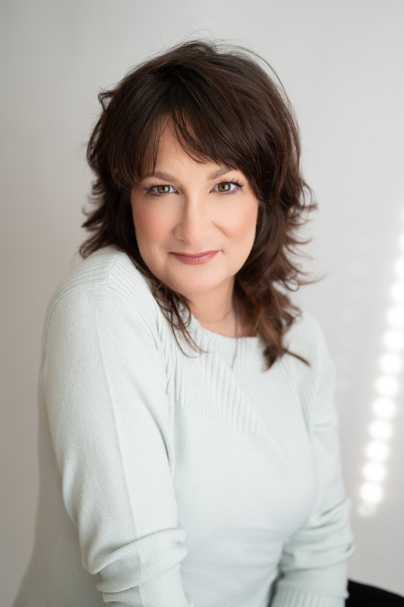 A woman with shoulder-length wavy brown hair and fair skin, wearing a white sweater, is smiling softly and looking directly at the camera with a neutral background.