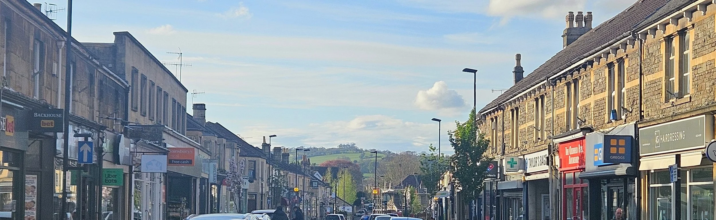 Moorland Road in Bath, a busy shopping street in Oldfield Park, featuring Oldfield Pharmacy offering NHS pharmacy services, travel vaccinations, and accessible parking.