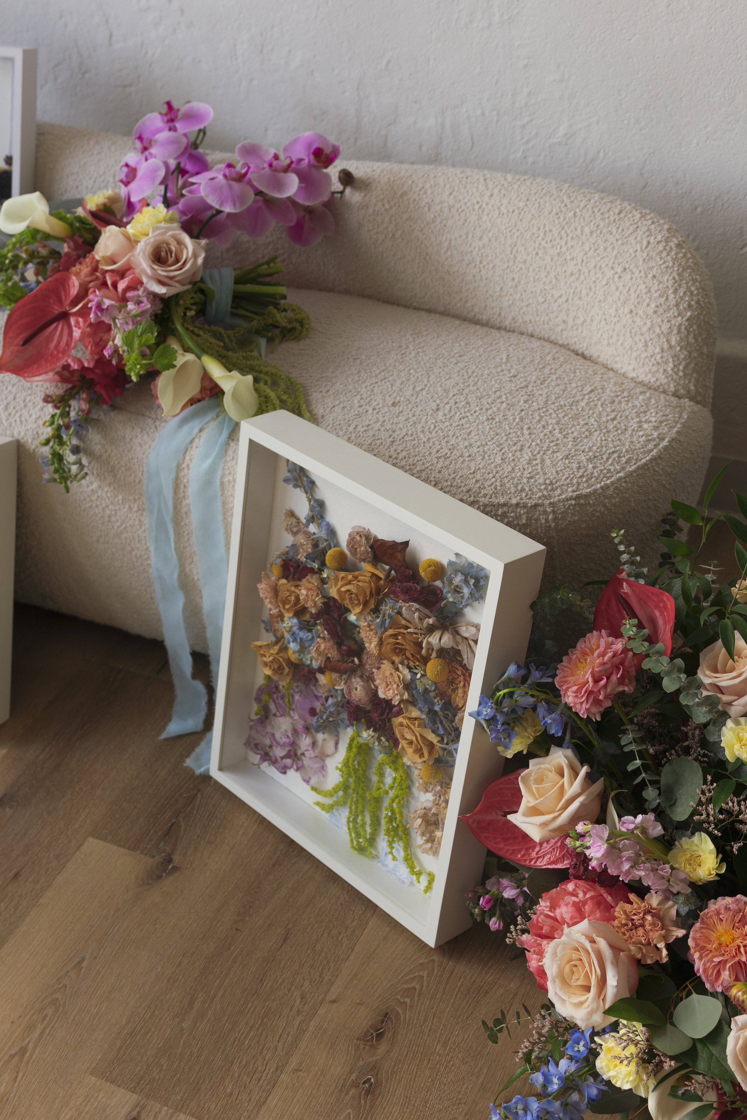 A floral arrangement with pink, yellow, cream, and blue flowers is placed on a beige textured sofa, with additional flowers scattered on the floor and a white shadow box filled with dried flowers nearby.