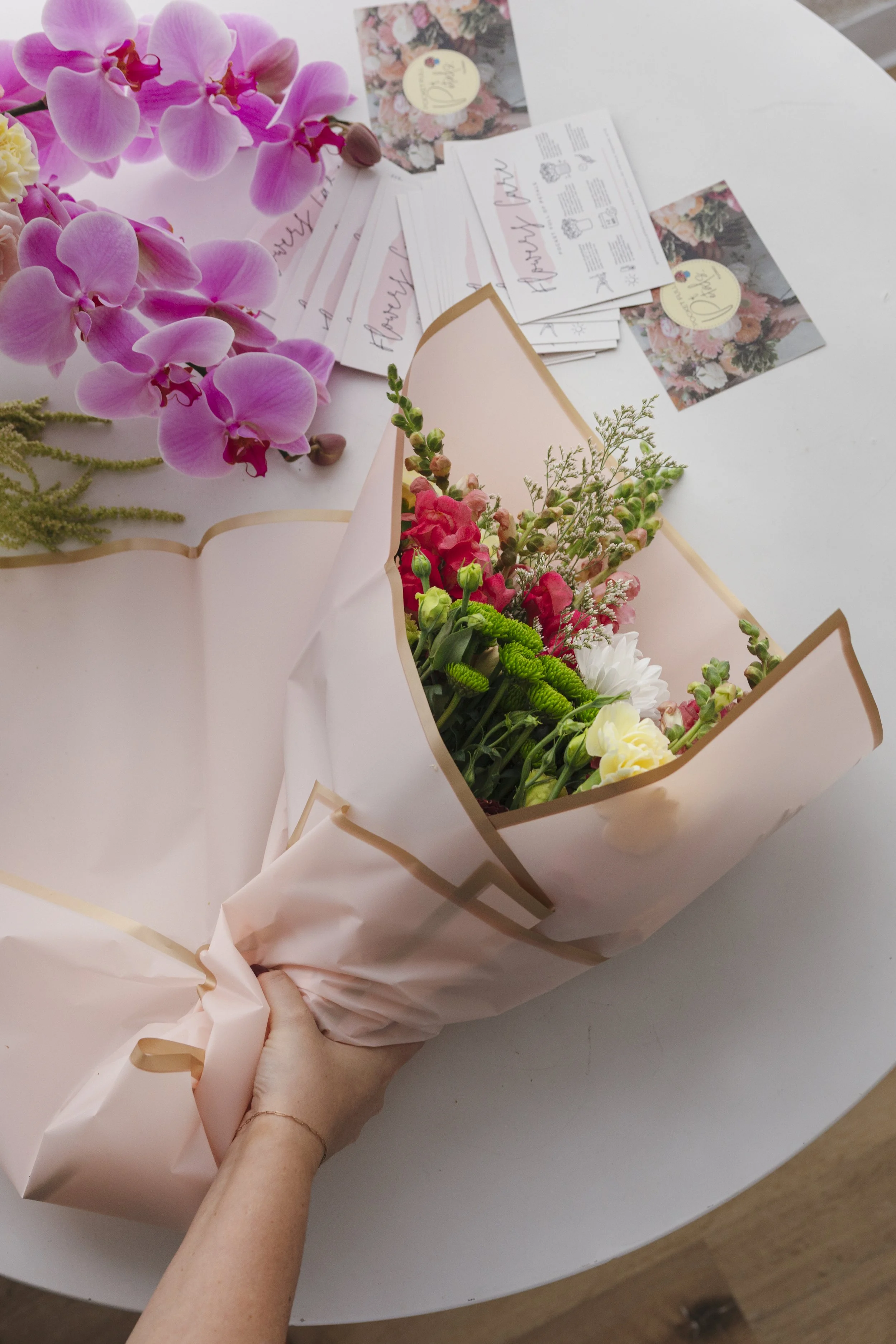 A hand holding a bouquet of pink, red, white, and green flowers wrapped in pale pink paper on a white table, with lavender and floral greeting cards in the background.