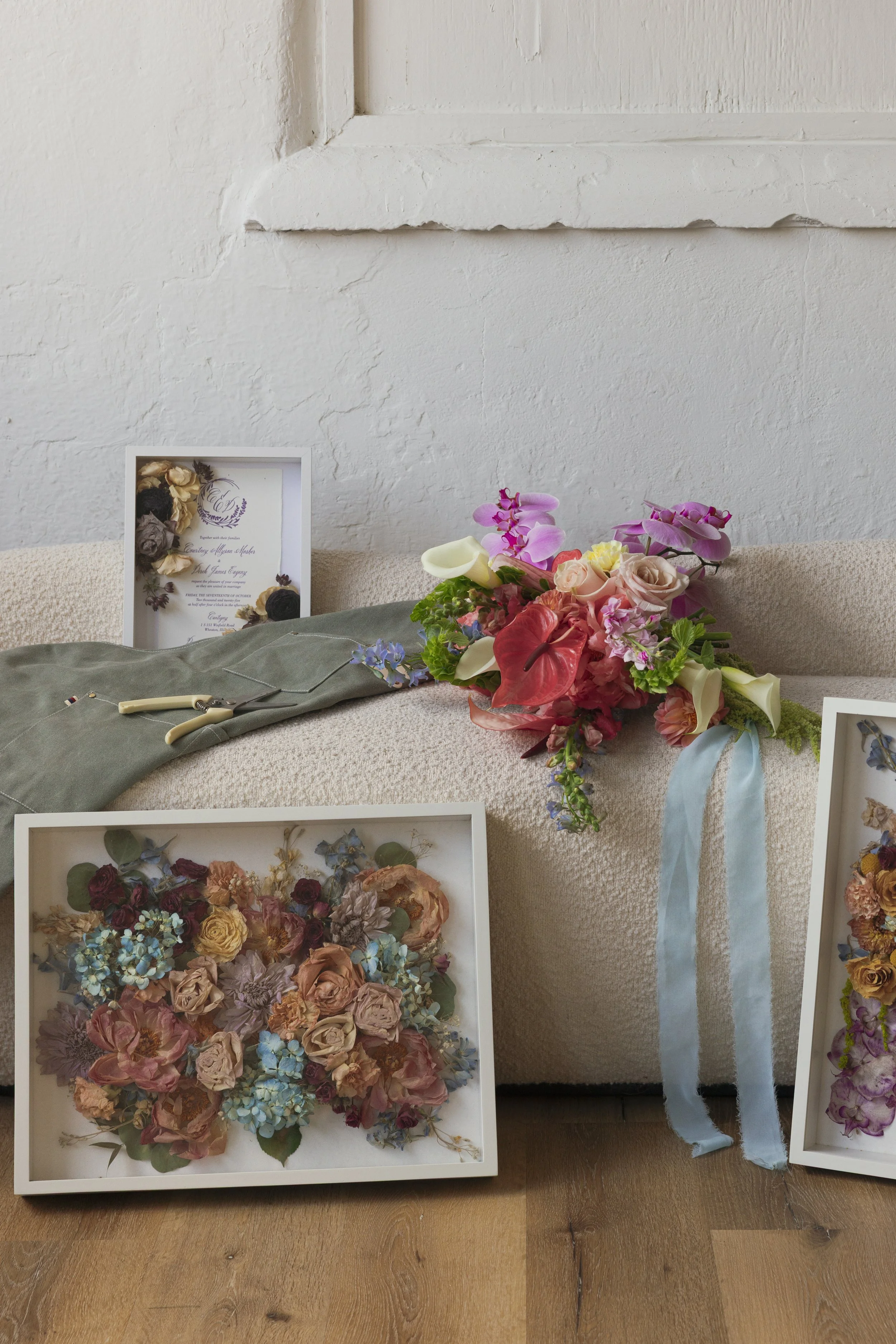 A bouquet of pink, white, and purple flowers with a blue ribbon on a beige couch, surrounded by framed floral artwork and a certificate.