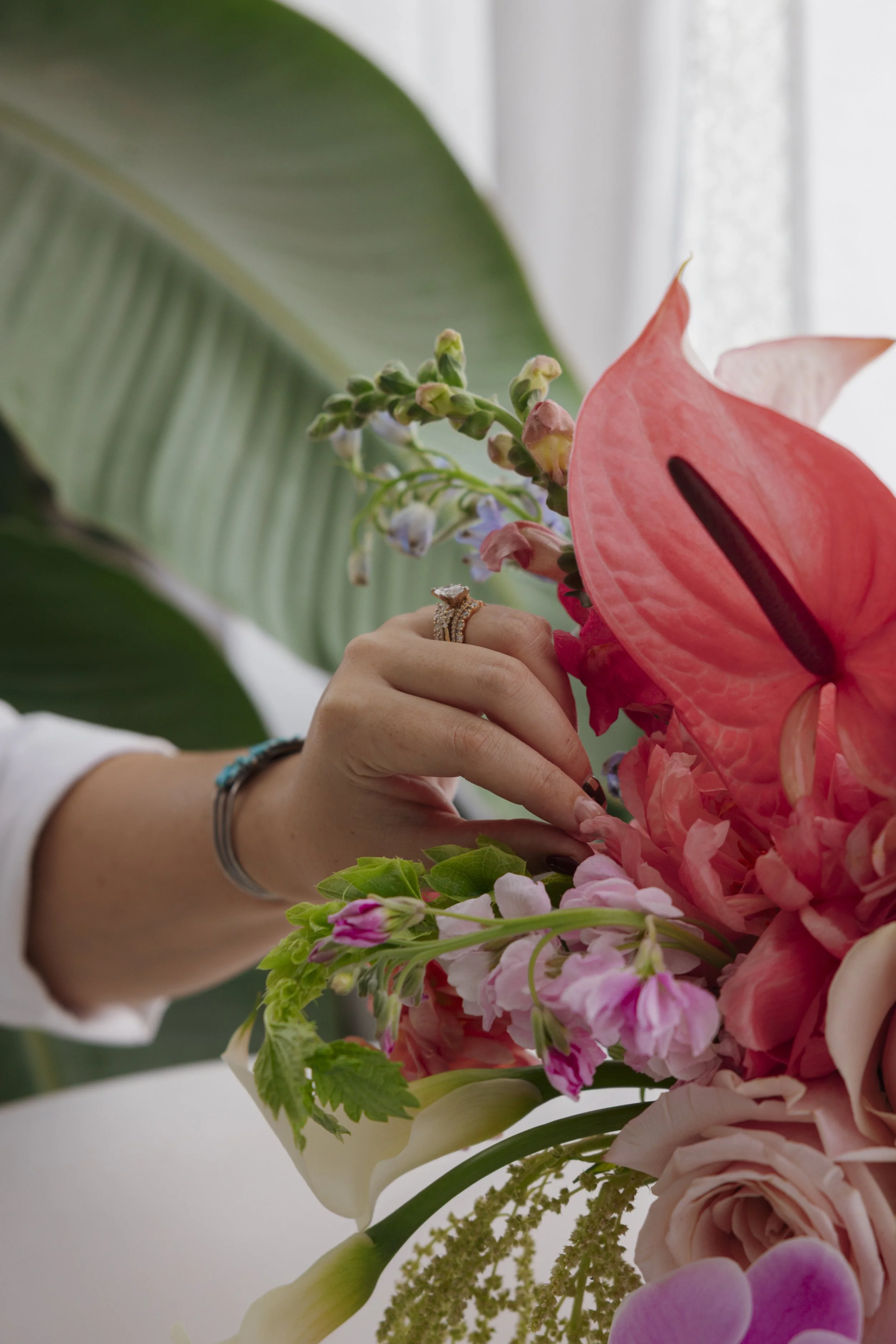 Close-up of a person's hand adjusting a bouquet of pink, red, and white flowers, including an anthurium, roses, and other blossoms, with large green leaves in the background.