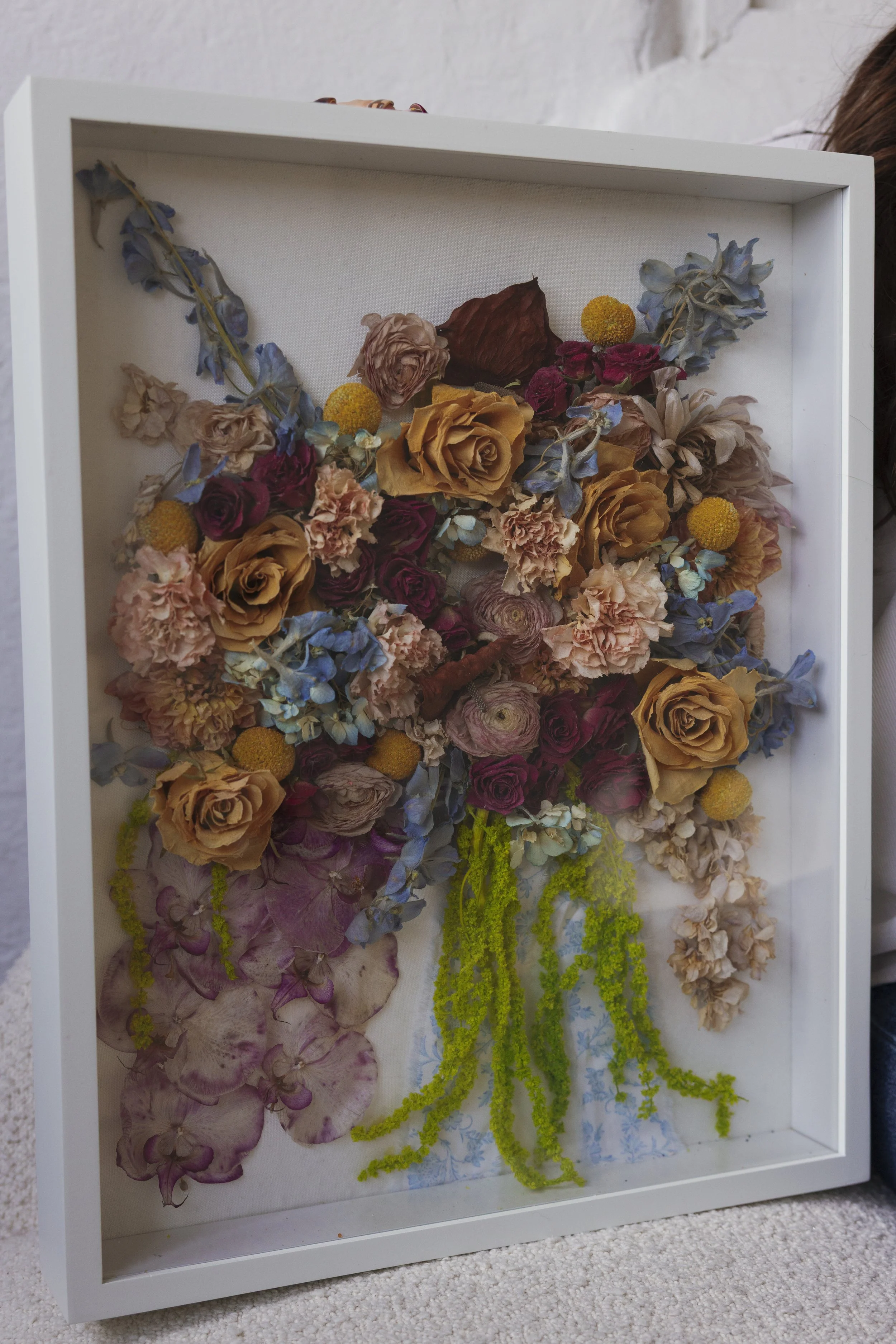 A framed display of dried flowers with various colors and types, including roses, hydrangeas, and other floral elements.