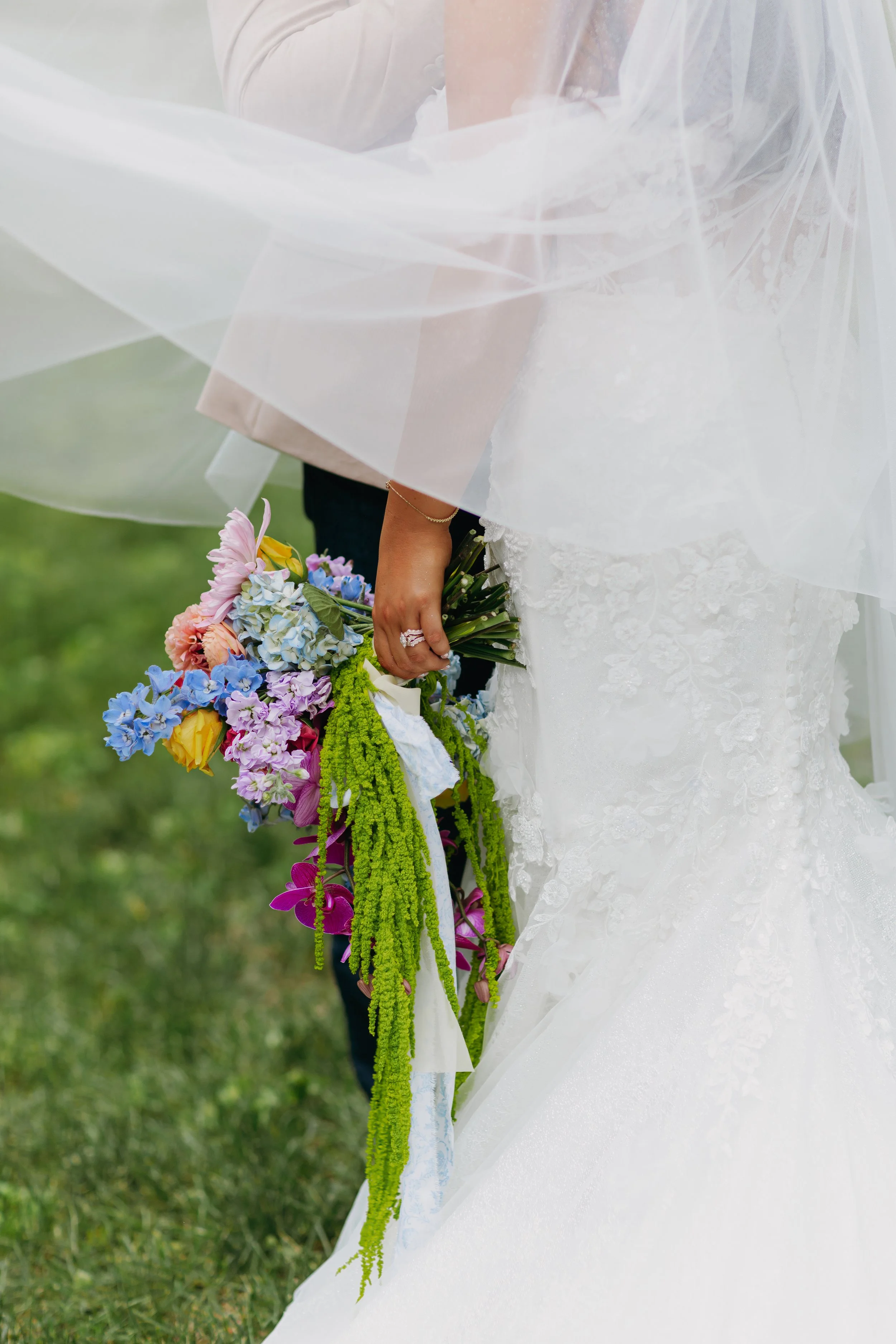 Bride holding a colorful bouquet of flowers, partially visible in her white wedding dress with lace details, outdoors on grass.