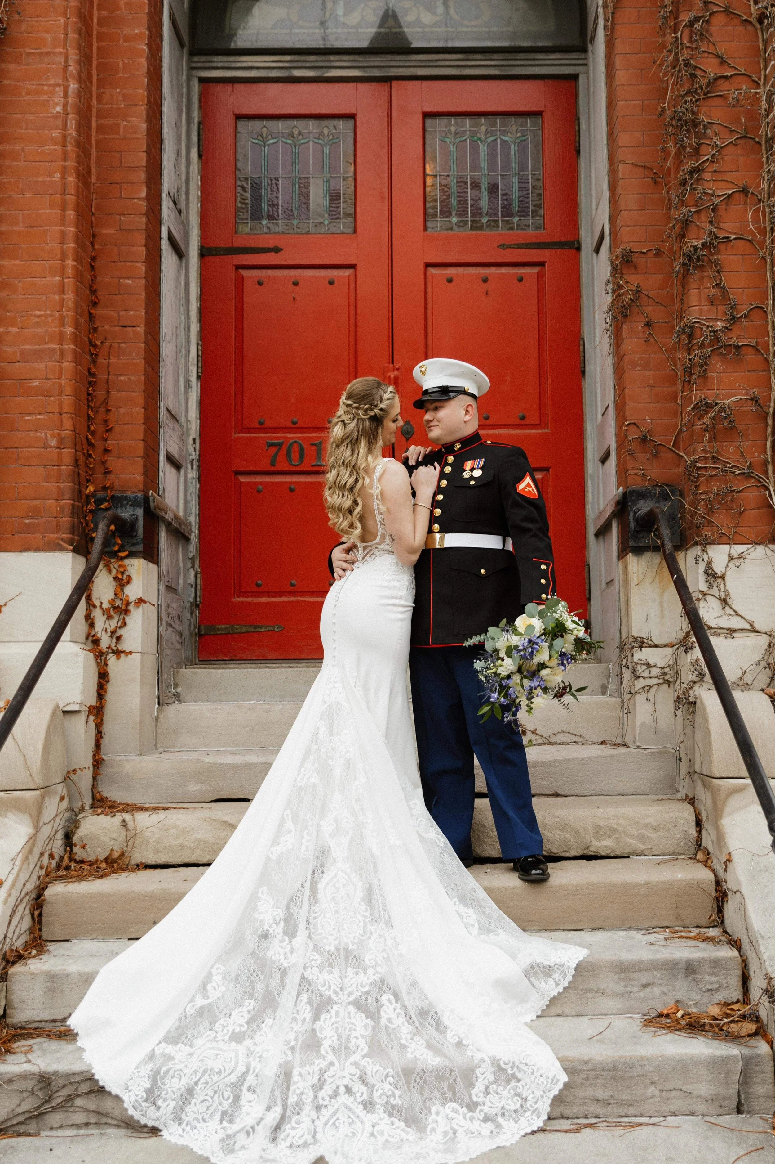 A bride and groom, with the bride in a white lace wedding gown and the groom in a U.S. Marine dress uniform, standing on steps in front of a red door, sharing an intimate moment.