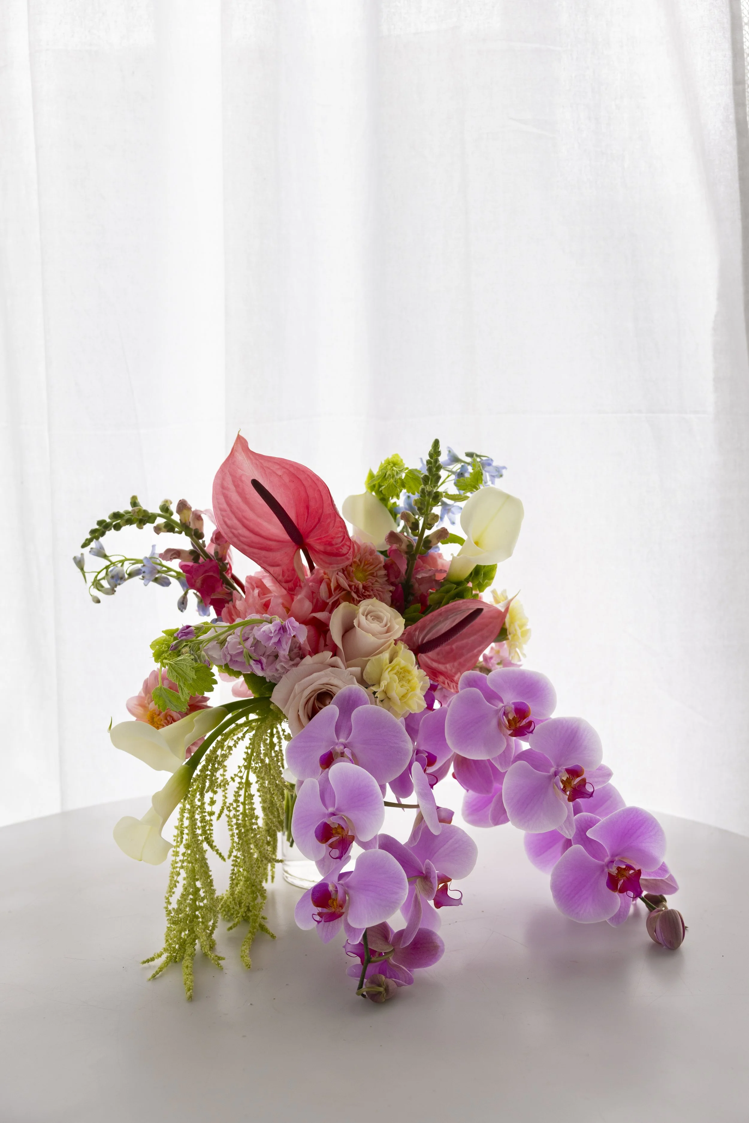 A colorful bouquet of various flowers, including pink, white, purple, and green blossoms, arranged in a clear glass vase on a white surface with a light, sheer curtain background.