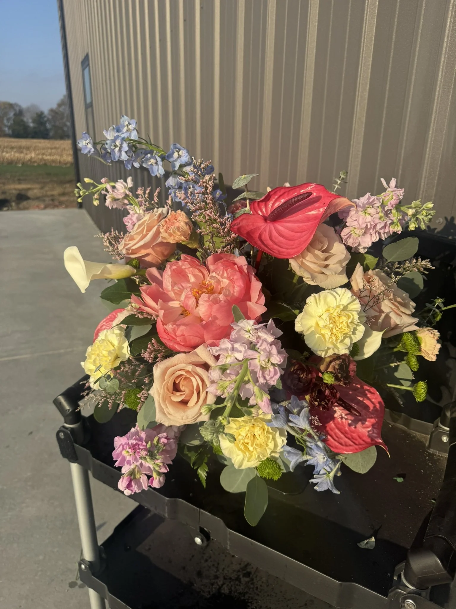 Colorful flower arrangement with pink, yellow, red, and purple flowers on a black cart outside, with a metal building and fields in the background.