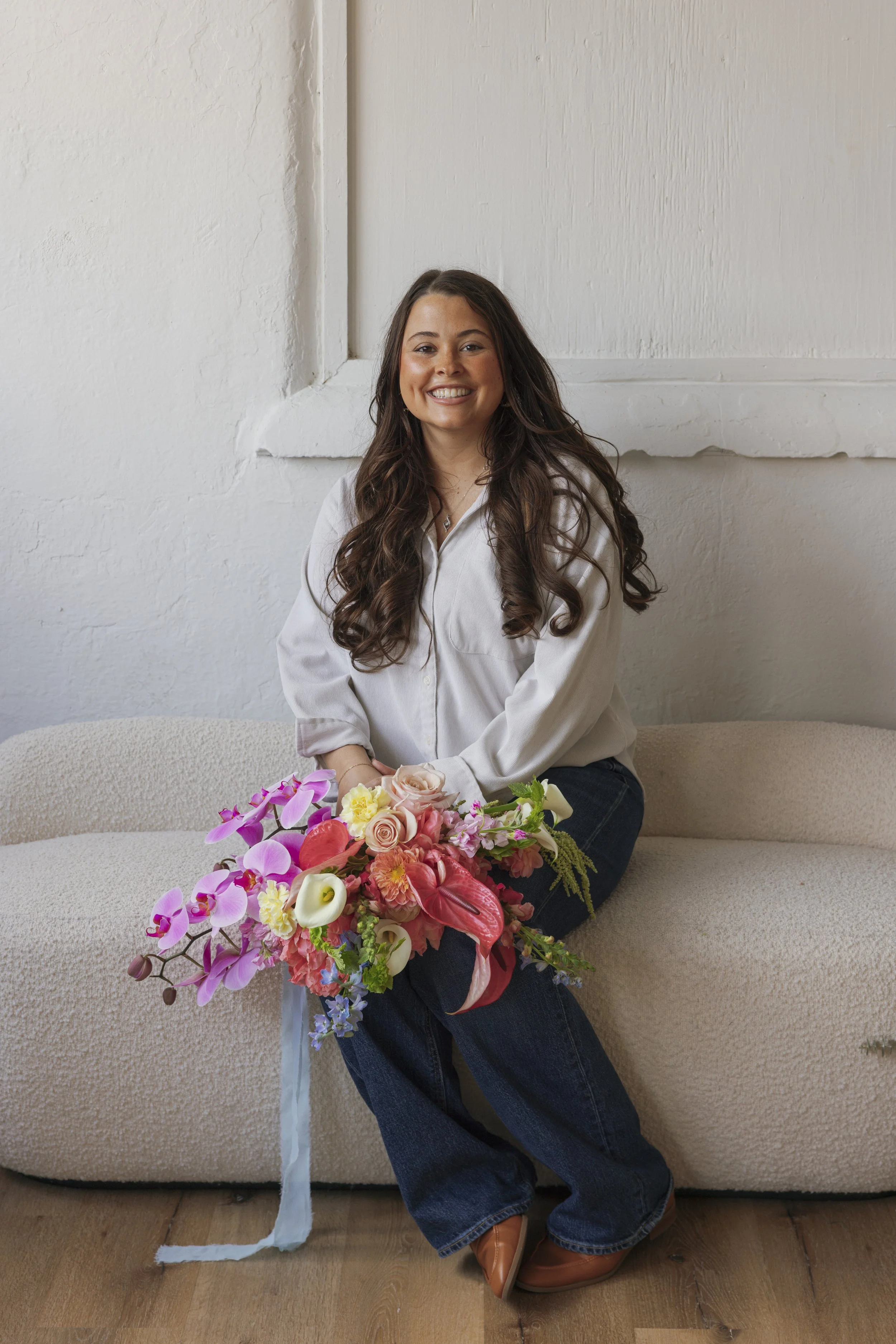 A woman with long curly brown hair sitting on a beige sofa, smiling, holding a large colorful flower bouquet.