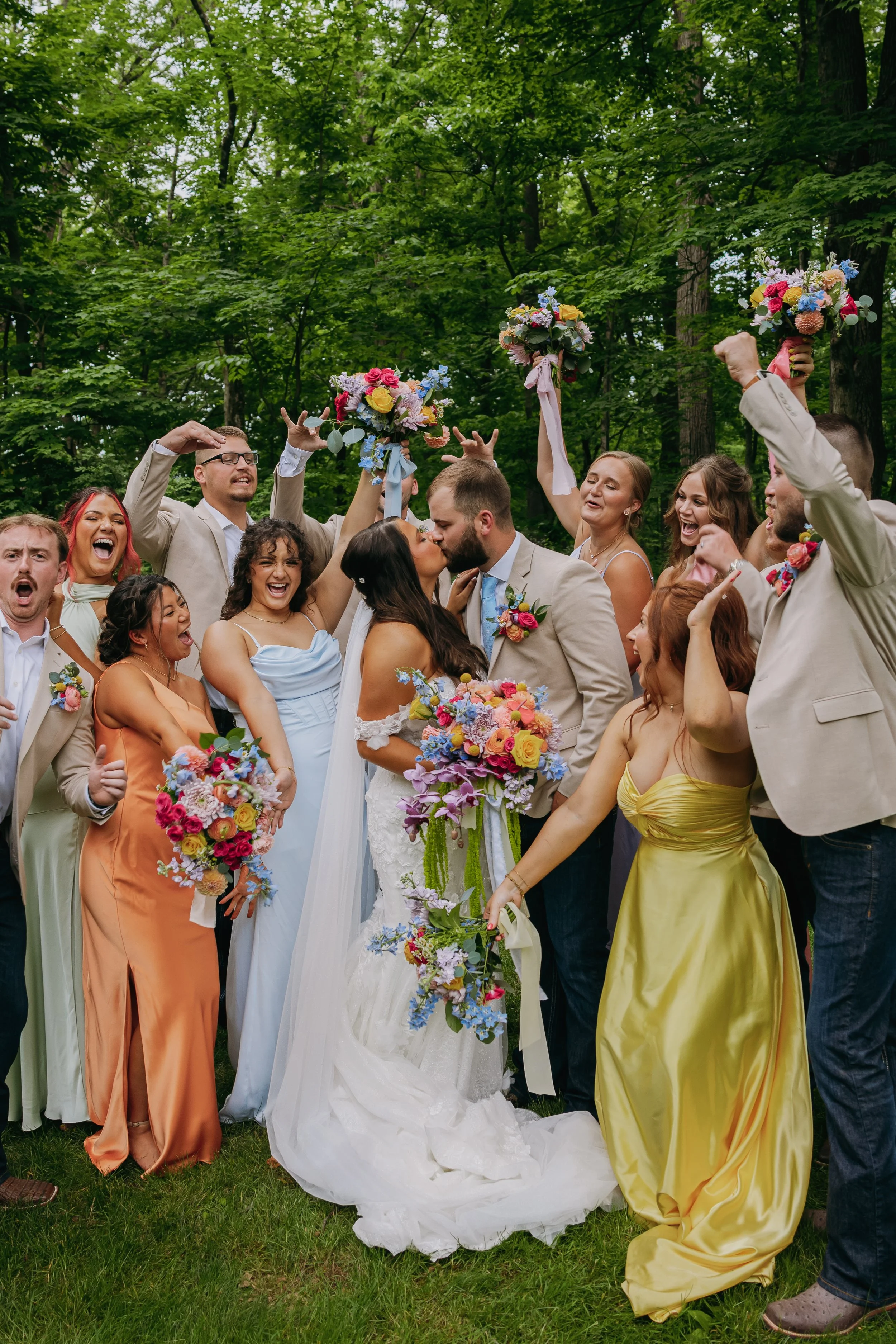 A wedding celebration outdoors with the bride and groom kissing surrounded by friends and family holding bouquets and celebrating.