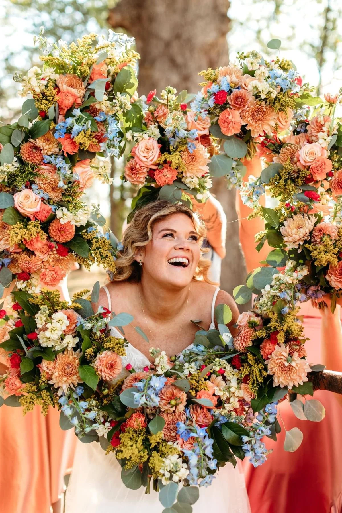 A woman with short blonde hair in a white dress is smiling through a large heart-shaped floral arrangement made of pink, orange, white, and blue flowers and green foliage. She is outdoors with trees in the background.