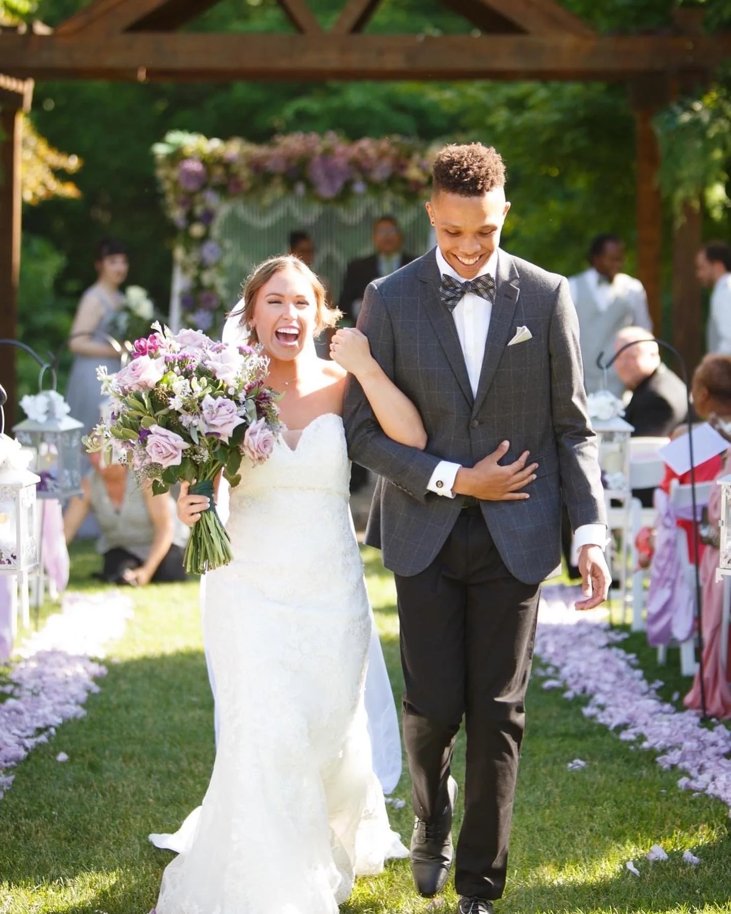 A bride and groom walking down the aisle at an outdoor wedding ceremony, surrounded by guests and greenery, with the bride holding a large bouquet of pink and purple flowers.
