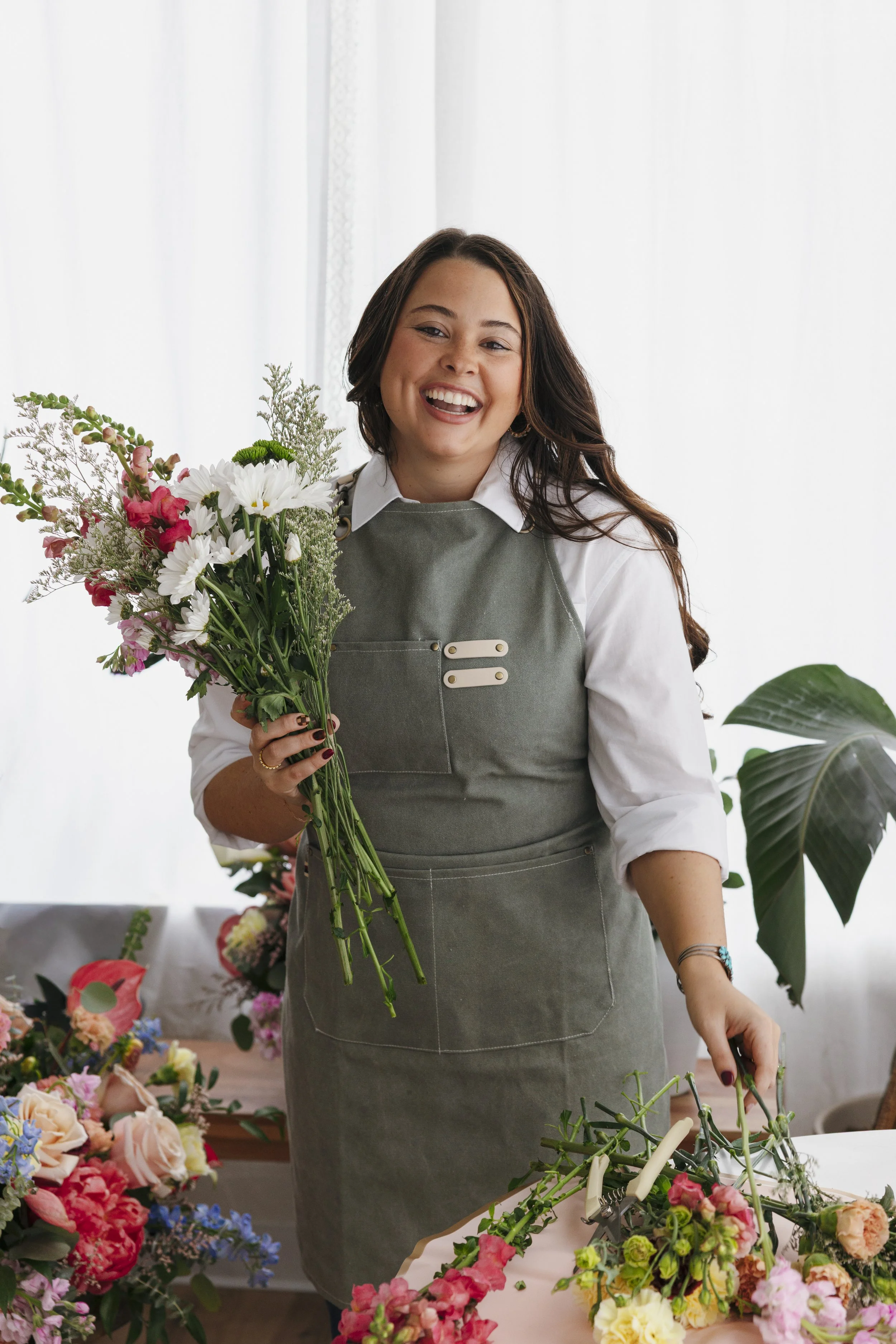 Woman smiling and holding a bouquet of mixed flowers, standing in a bright room with a table full of colorful flower arrangements.