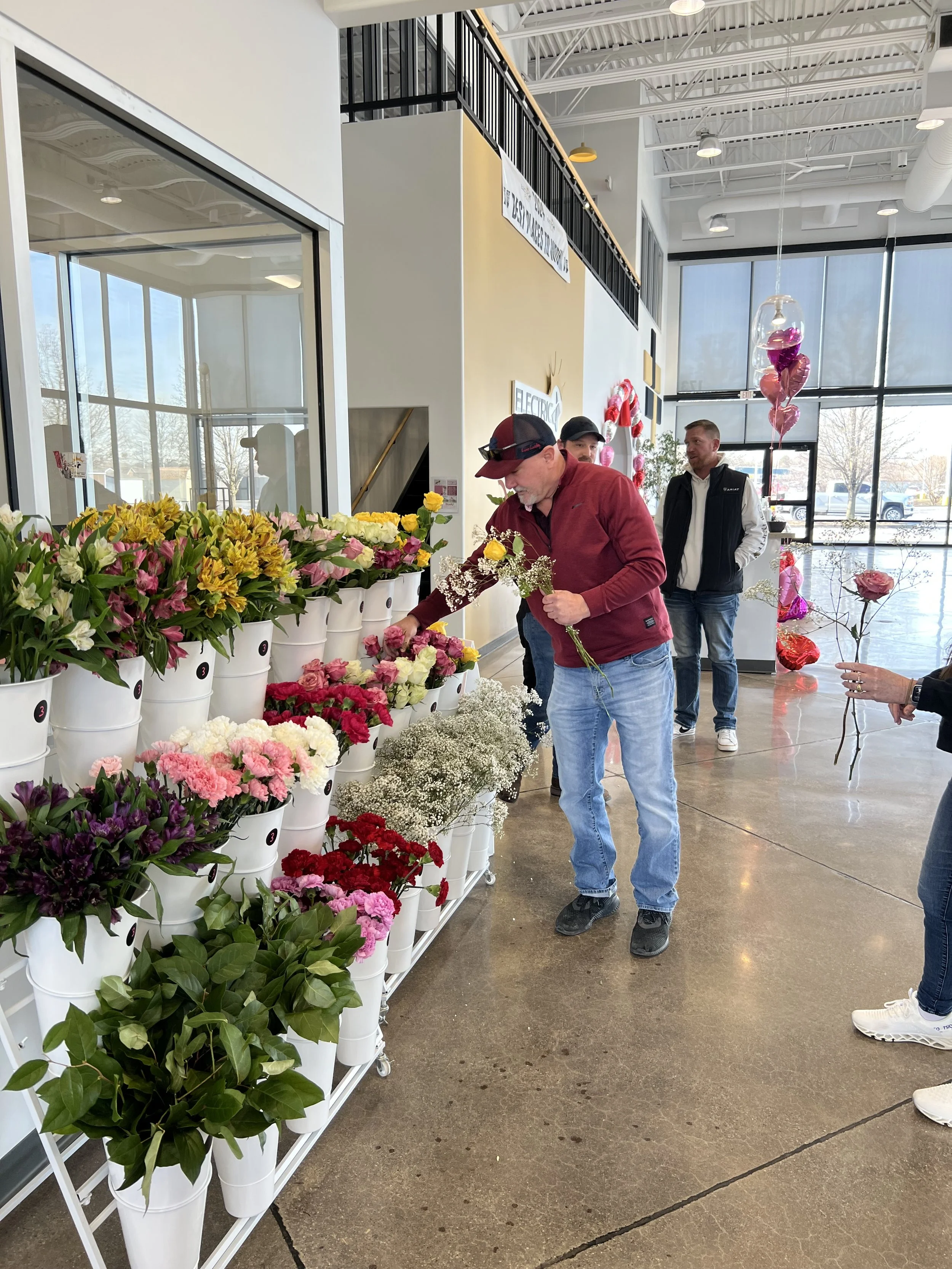 Man selecting flowers from a display of colorful flowers in a store, with people and Valentine's Day balloons in the background.