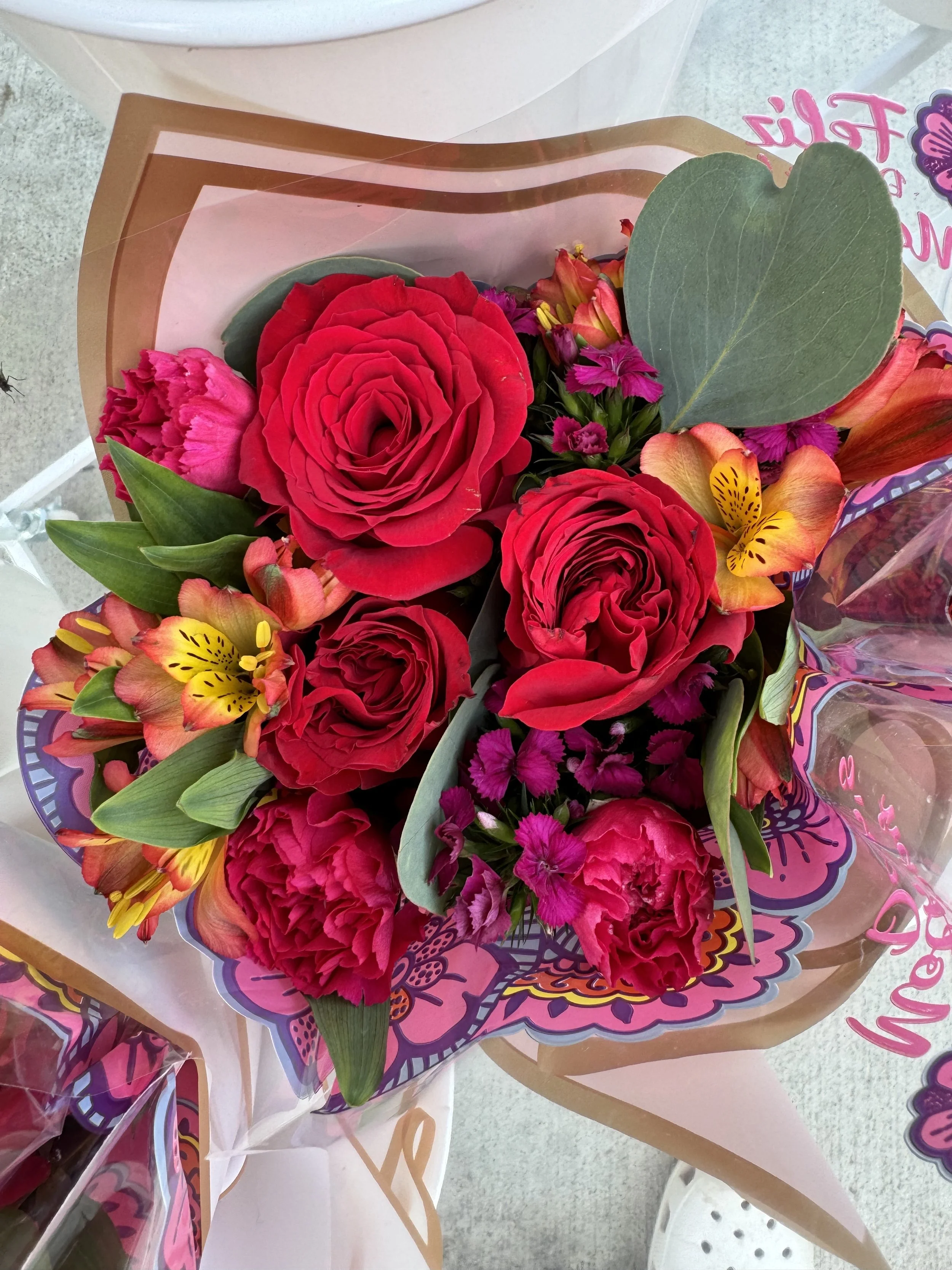 Bouquet of red roses, pink and yellow lilies, and small purple flowers wrapped in pink and brown paper.