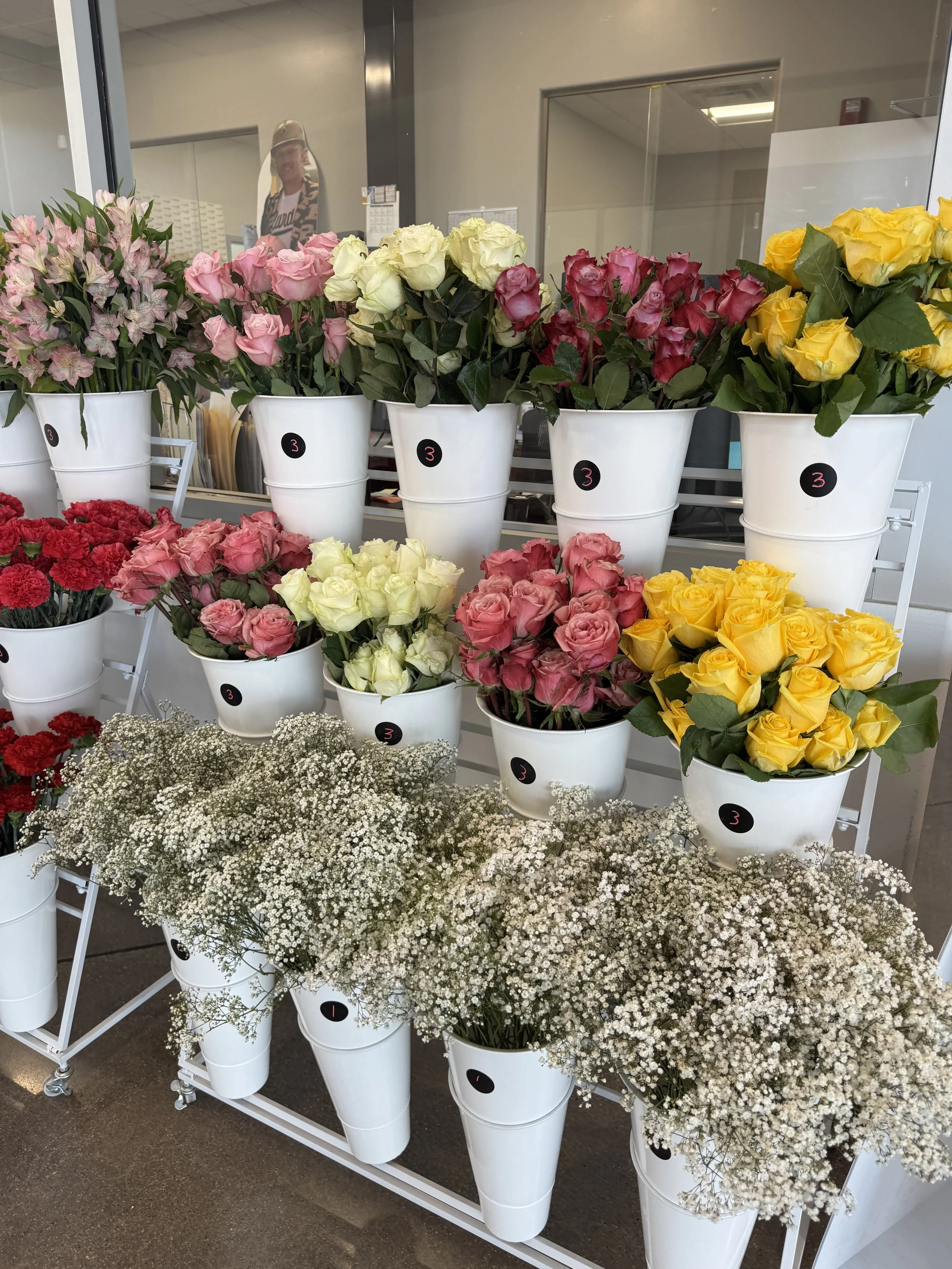 Display of various colorful flowers in white buckets at a flower shop, including pink, white, yellow roses, red carnations, and white baby's breath.