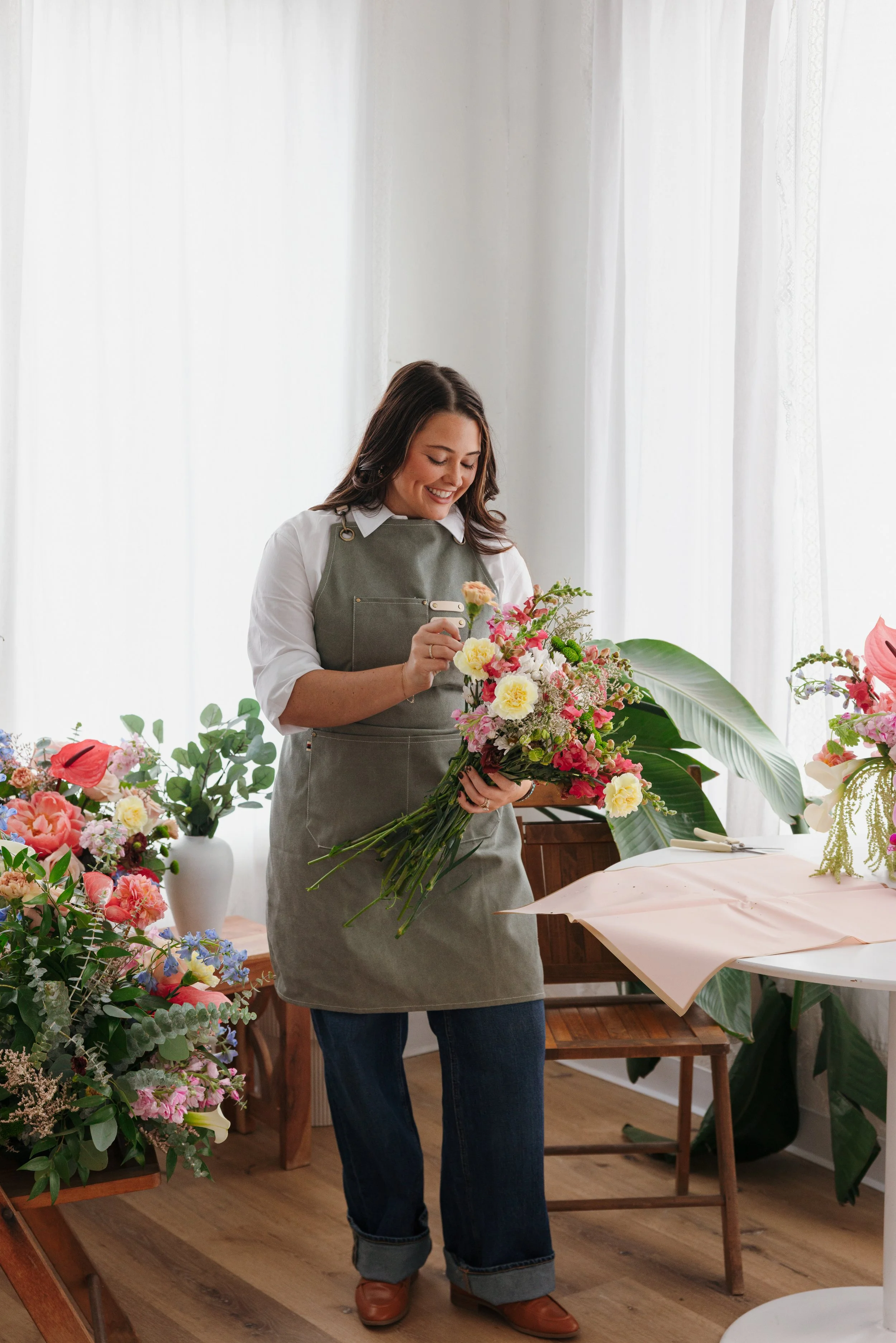 A woman arranging a bouquet of colorful flowers in a bright room with white curtains, wooden flooring, and plants.